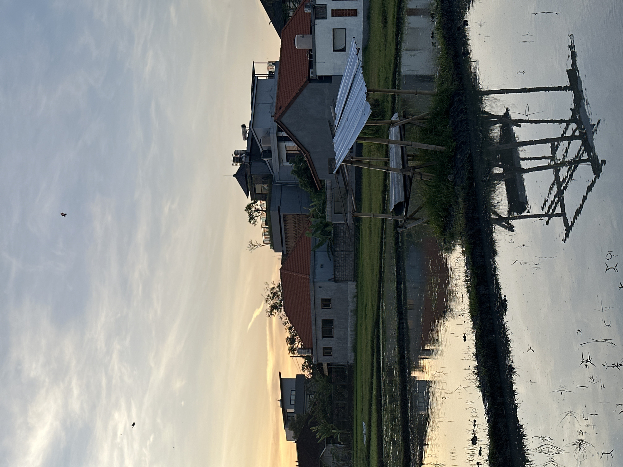 The image depicts a tranquil rural scene, likely captured in North Kuta, Indonesia, judging by the landscape and architecture. The scene focuses on a rice paddy field that reflects the sky and surrounding structures. Several modern buildings are on the edge of the rice field, with a variety of roof designs. A small bamboo structure with a corrugated metal roof stands near the edge of the paddy, its reflection visible in the water. The sky transitions from a bright yellow-orange near the horizon to a light blue with scattered clouds above. There appear to be some birds or kites in the sky, adding to the outdoor atmosphere. The reflection in the water captures the sky, buildings, and structure, creating a mirrored effect that enhances the visual depth of the landscape.