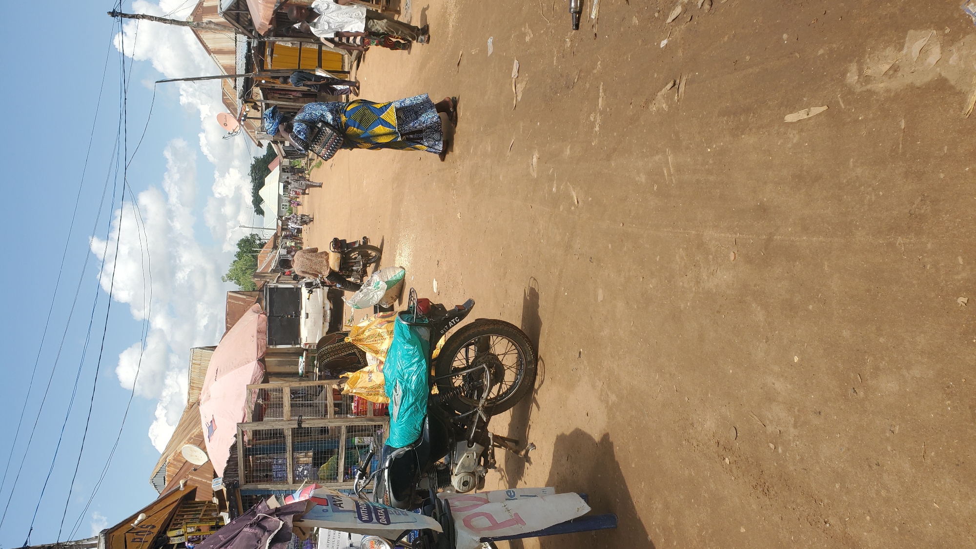 The image depicts a dusty, unpaved street in Jalingo, Nigeria, under a bright, partly cloudy sky. The street is lined with small shops and market stalls constructed from corrugated metal and wood. Several people are present, engaged in various activities. In the foreground, a black motorcycle with a turquoise cover over its seat is parked. To the left, a market stall displays goods, including bags of rice. Further down the street, a woman in a blue and yellow patterned dress stands with a bag on her shoulder. Other individuals are seen walking or riding motorcycles, indicating ongoing market activity. The environment is characterized by its rural, open-air market setting.