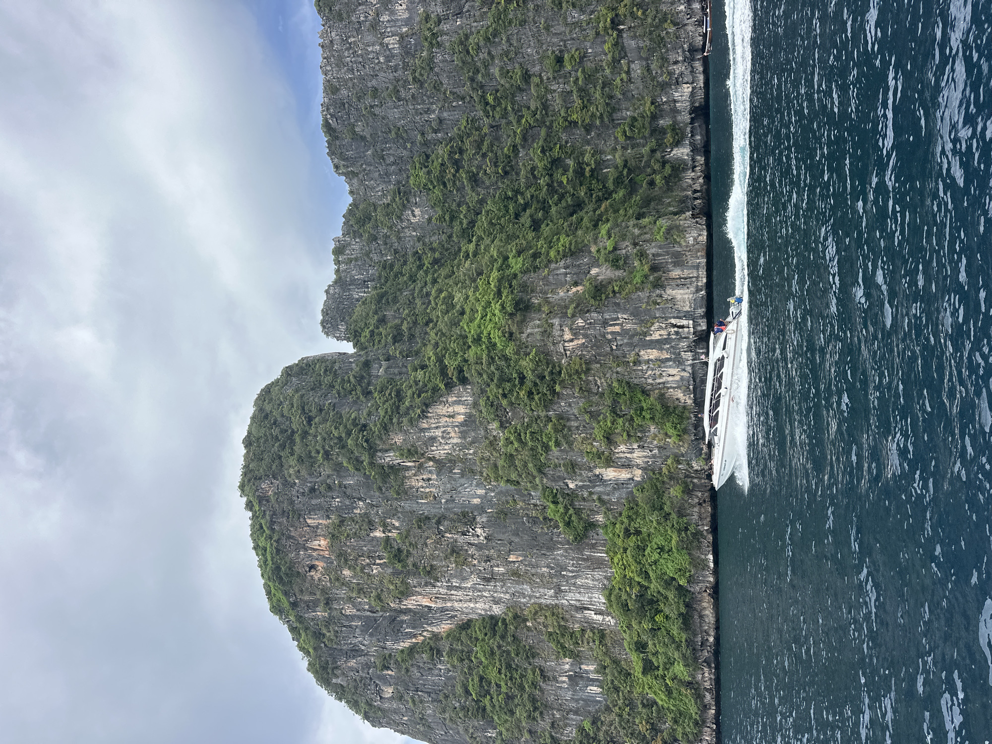 A high-angle, full shot captures a speedboat moving to the left on dark blue water. The boat is white with a dark stripe and has several passengers visible. Behind the boat, a steep, rocky cliff face covered in lush green vegetation rises dramatically from the water. The cliff has vertical striations and appears weathered. The sky above is overcast with gray clouds. The scene is located at Haad Hin Khom, Thailand.