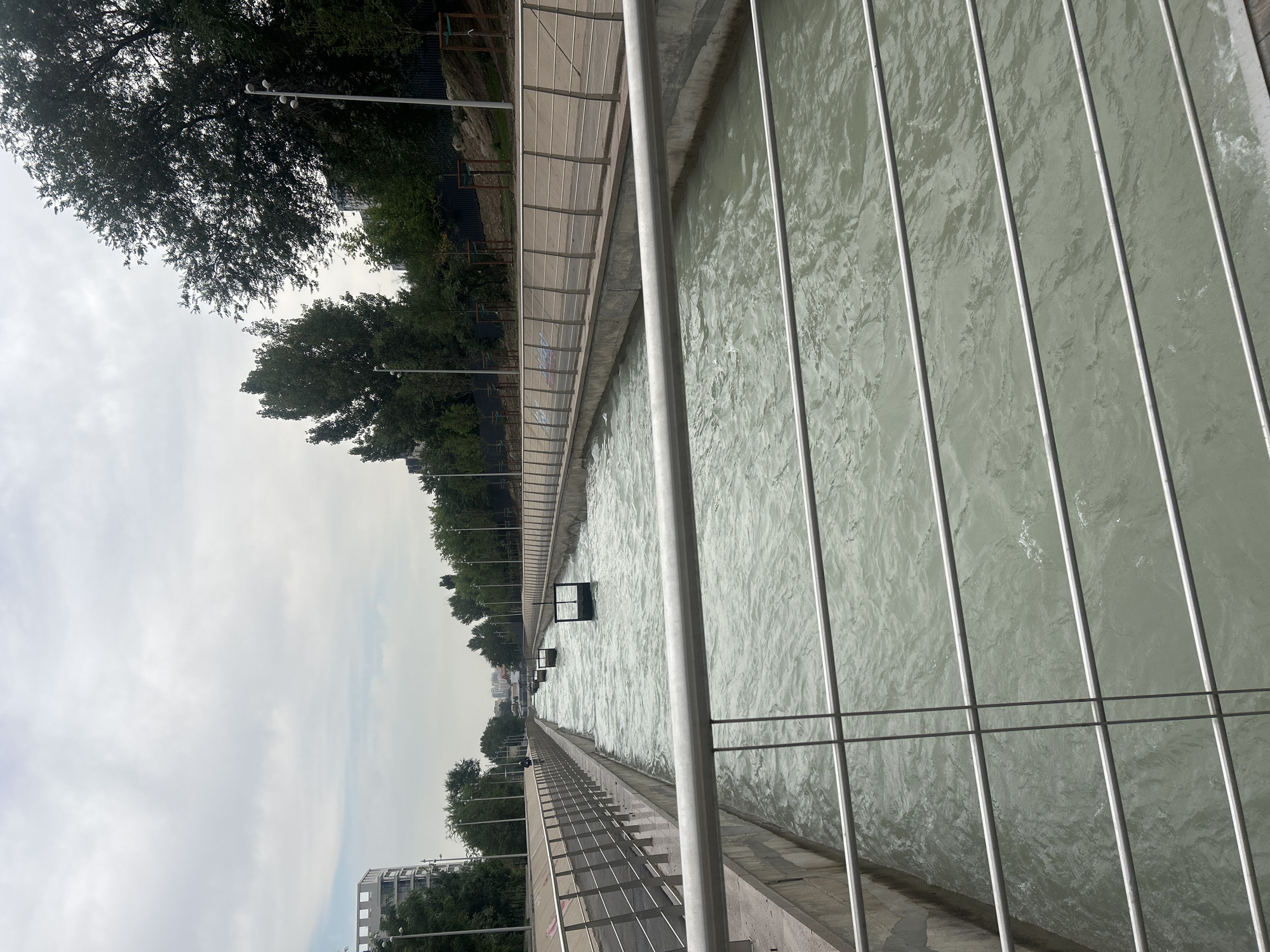 The image depicts a man-made canal with flowing water, likely part of an urban development project in Almaty, Kazakhstan. The canal is lined with concrete walls and has metal railings along its sides, suggesting a pedestrian walkway or public space. Several square, dark-colored structures, possibly water control gates or weirs, are visible within the canal. Trees and buildings are present on both sides of the canal, with a cloudy sky overhead. The water appears somewhat murky and agitated, indicating movement. In the distance, buildings and more trees are visible, hinting at a developed urban environment. The overall scene suggests a recreational or functional waterway within a city.