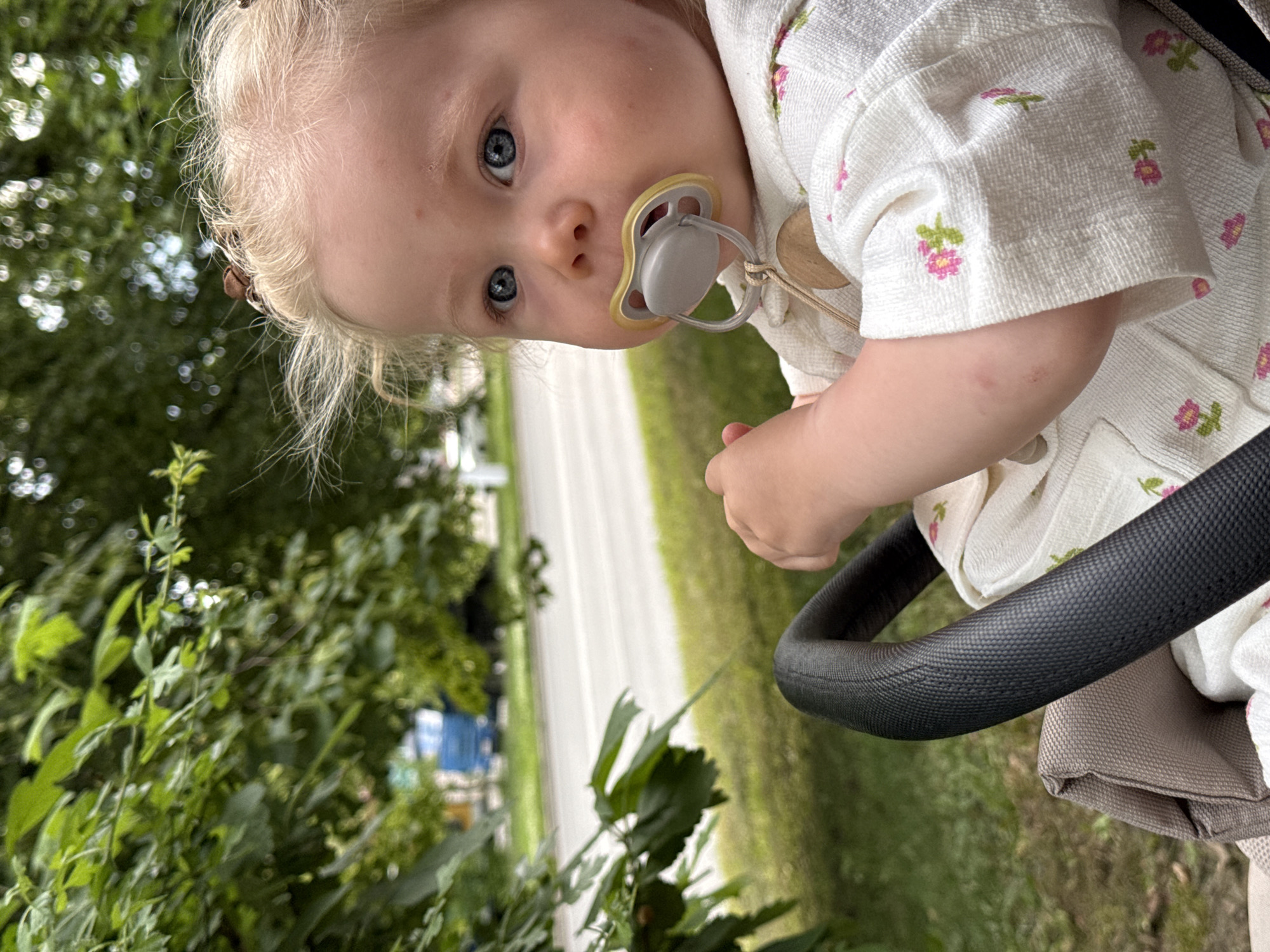 A close-up, low-angle shot captures a baby girl with blonde hair and blue eyes, seated in a stroller. She has a pacifier in her mouth and is wearing a white, floral-patterned garment. The background consists of blurred green foliage and a paved road with some distant vehicles, suggesting an outdoor urban setting. The lighting is natural, indicating it is daytime. The stroller's black handle is visible in the foreground. The scene does not provide specific landmarks to identify a location within Vladimir, Russia.