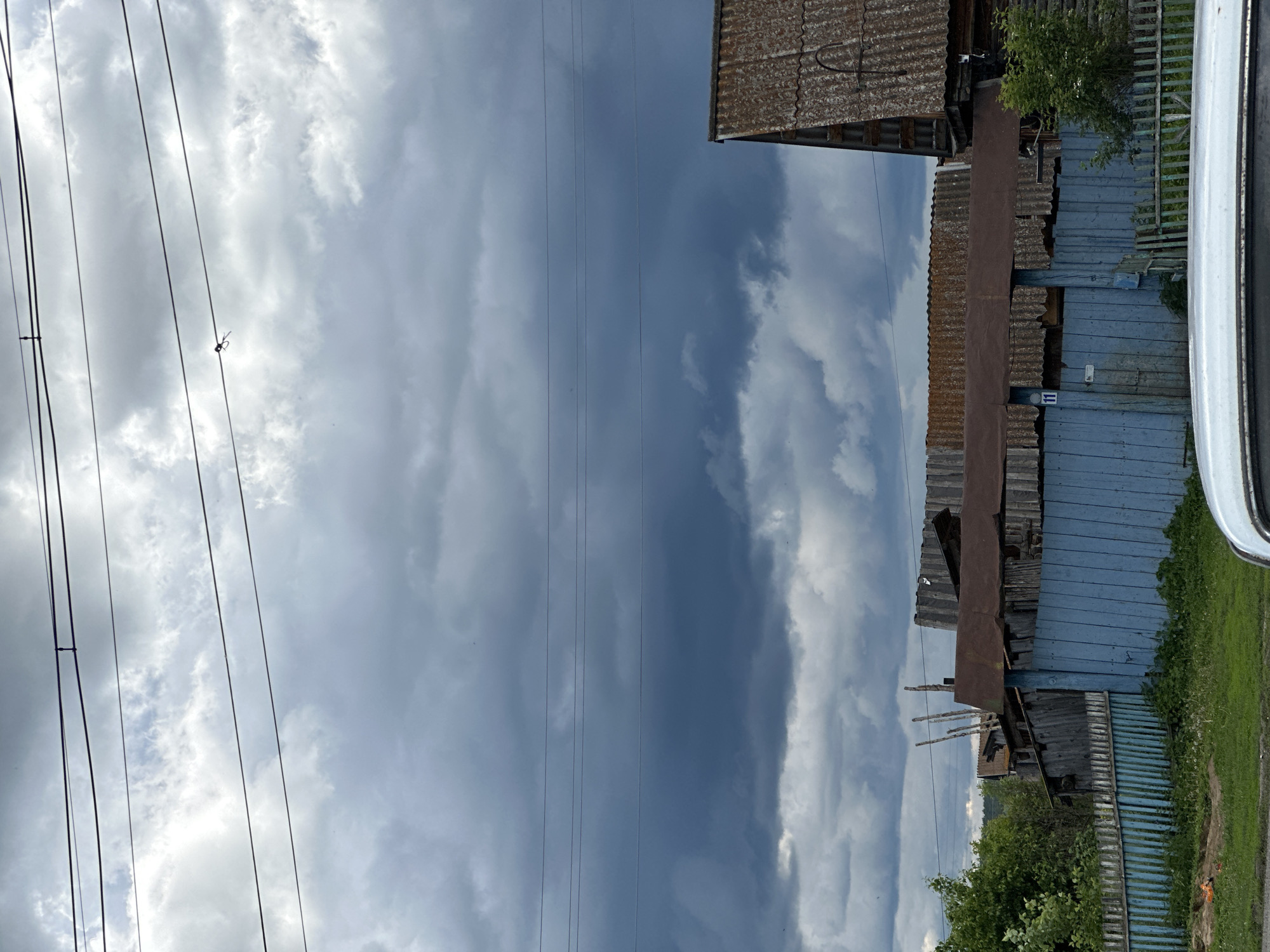 The image depicts an outdoor scene with a dramatic, cloudy sky. Several dark, heavy clouds dominate the upper portion of the frame, with lighter, wispy clouds interspersed. Multiple power lines stretch horizontally across the sky. Below the sky, there is a collection of rustic buildings and fences. A prominent blue wooden fence runs horizontally across the lower part of the image, with a section of a lighter, green wooden fence visible to the right. Behind the blue fence, there are structures with corrugated metal and wooden roofs, some appearing weathered and aged. Trees and dense foliage are visible to the left of the fence line. The foreground shows a patch of green grass and the edge of a white vehicle. The overall impression is of a rural or semi-rural setting under a stormy sky.