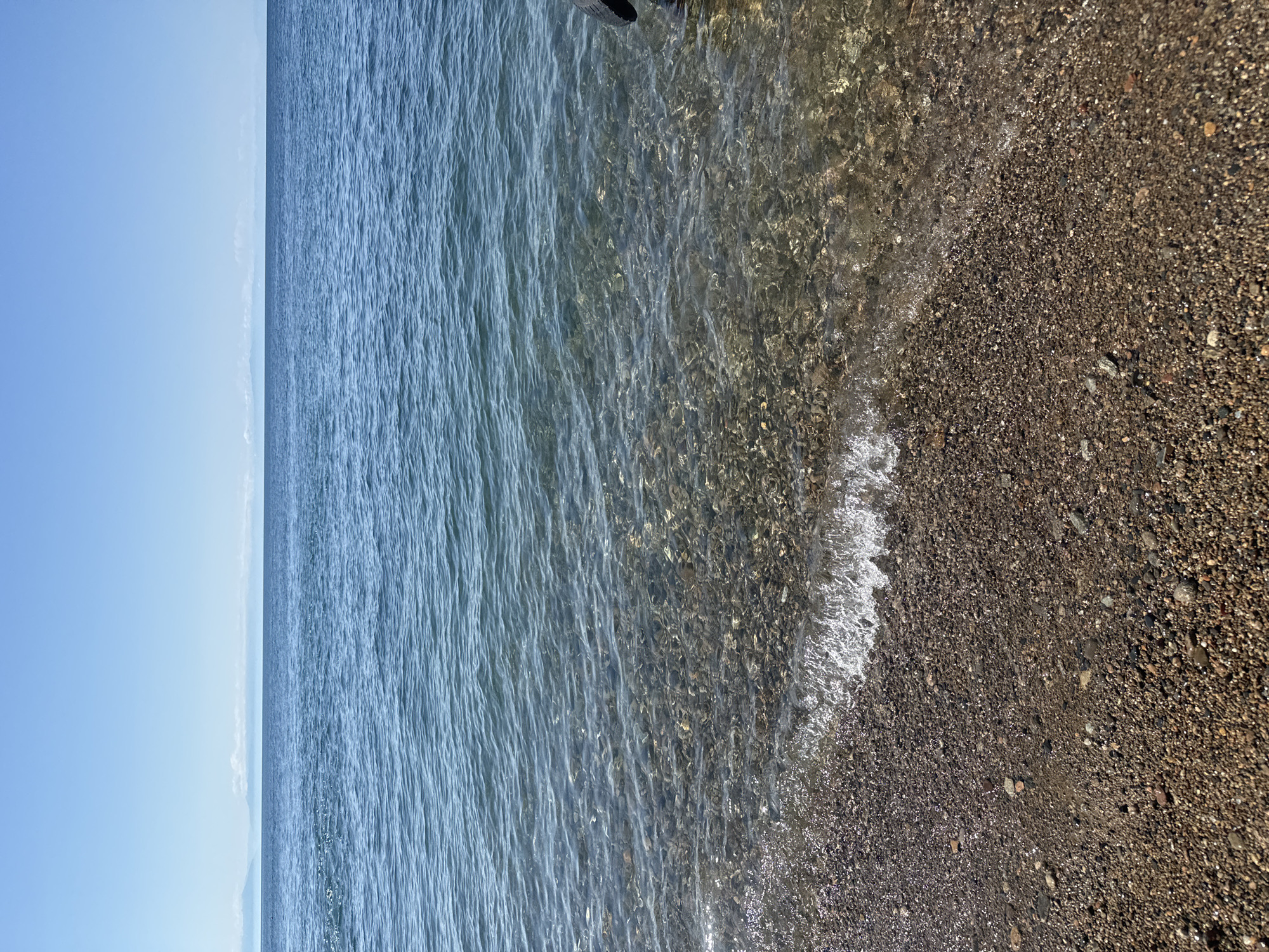 The image depicts a tranquil scene at the shore of Lake Baikal in Listvyanka, Russia. The foreground is a pebbled beach, wet from the receding wave that breaks with white foam. The water is clear, allowing a view of the submerged stones on the lakebed. Beyond the shallows, the lake stretches out, its surface rippled by a gentle breeze. In the distance, a hazy line of mountains under a clear blue sky marks the horizon. A small portion of a dark object, possibly a tire or part of a boat, is visible on the right edge of the frame. The overall impression is one of natural beauty and calm.