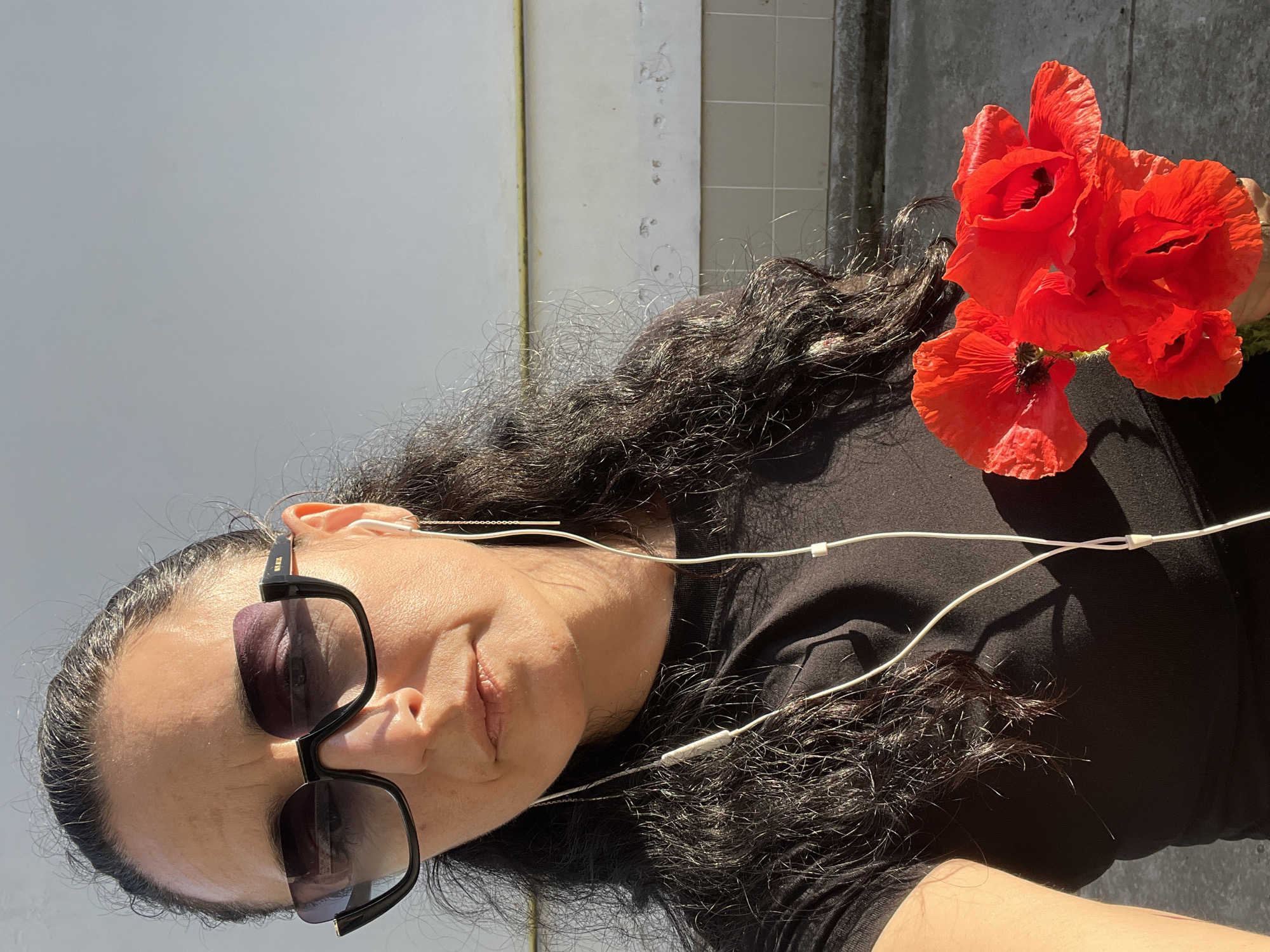 A middle-aged woman is depicted in a selfie-style shot in Tarnivtsi, Ukraine. She has dark, wavy hair pulled back and wears oversized black sunglasses with gradient lenses, featuring the brand "MARQON" on the arm and "M.M." on the temple. White wired earbuds are in her ears, with the wire visible draped across her black short-sleeved top. A gold-colored ear threader earring is present in her right ear. She holds a bouquet of approximately five vibrant red poppy flowers with dark centers in her left hand, positioned in the lower right foreground. Her expression is neutral, and she looks directly towards the camera. The scene is outdoors under direct sunlight, casting distinct shadows on her face, neck, and the flowers. The background consists of a light-colored, textured wall with visible imperfections. A horizontal yellow pipe traverses the mid-section of the wall, below which is a band of light beige square tiles. The ground surface is dark gray asphalt or concrete.