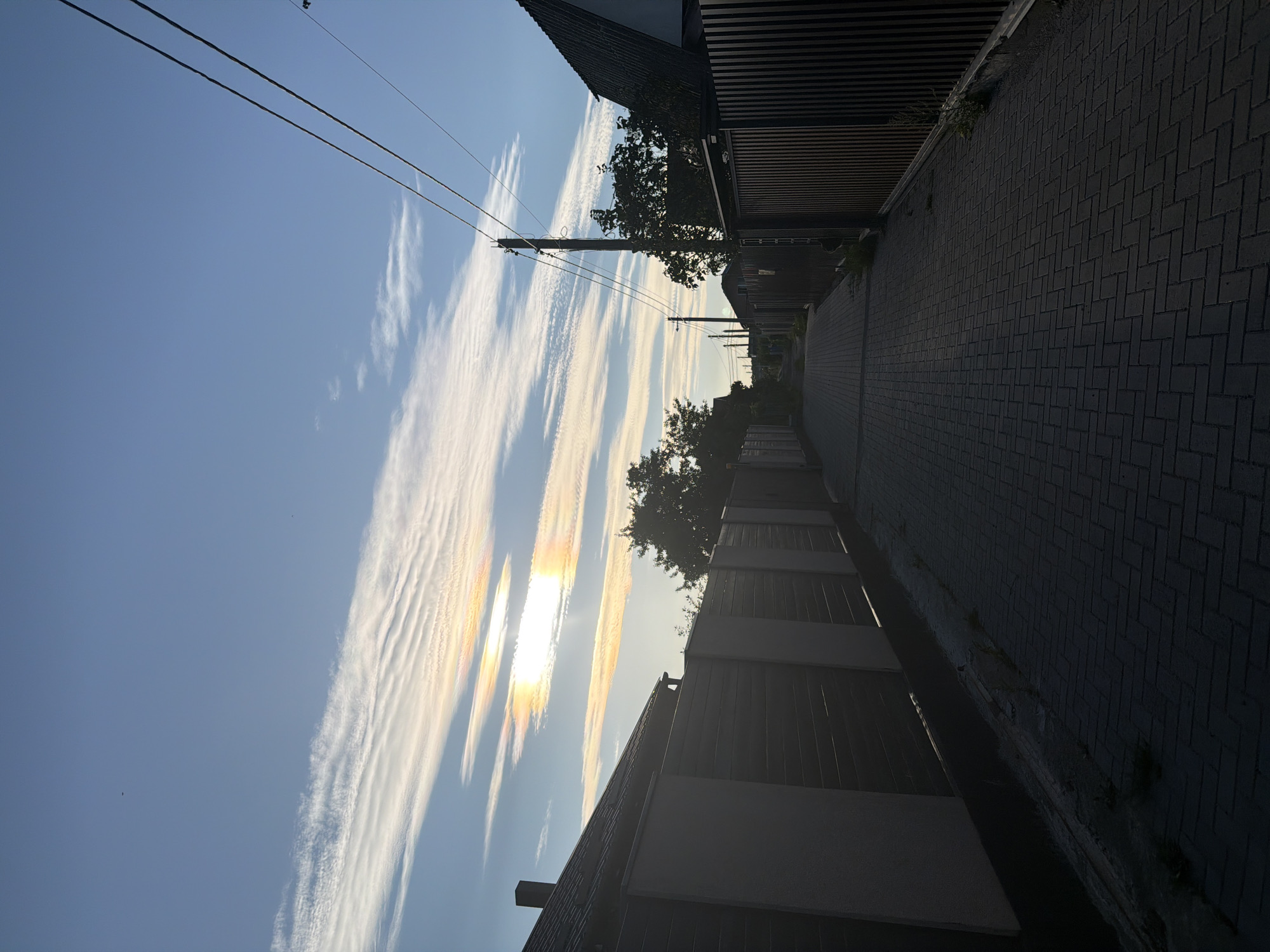 This is an eye-level, full shot of a paved pathway leading into the distance between fences on either side. The sky above is a clear blue with some streaky, wispy clouds that exhibit rainbow-like iridescence. On the left, a dark wooden fence with lighter colored vertical posts runs along the pathway. Behind the fence are the dark roofs of houses. On the right, a similar dark wooden fence is visible, with a wooden utility pole and power lines overhead. Trees are visible behind the fences in the distance. The pathway is paved with dark gray interlocking bricks laid in a herringbone pattern. The lighting suggests it is either early morning or late afternoon, with shadows cast by the fences and houses. There are no people or animals visible in the frame. The scene appears to be a residential street or alleyway in a city.
