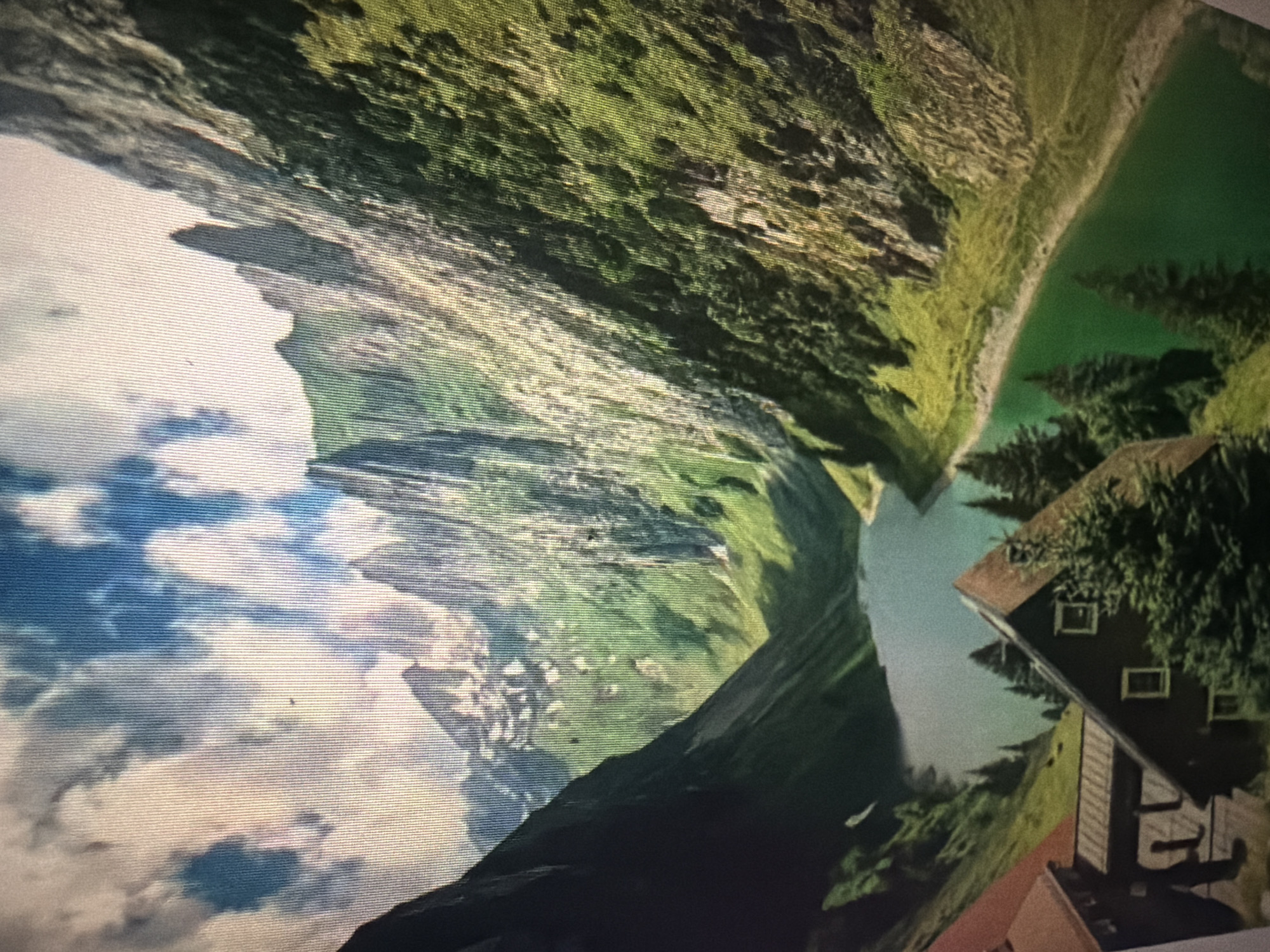 This media file depicts a surreal, inverted landscape with a dramatic mountainous backdrop and a body of water. The scene is dominated by lush green, grass-covered slopes and jagged rocky outcrops, suggesting a mountainous region. In the foreground, to the right, a cluster of houses with dark roofs and visible windows is nestled amongst dark green trees. The sky, filled with fluffy white clouds, appears in the upper left portion of the image, but the overall orientation suggests the sky is actually below the landscape. The water is a vibrant, almost unnatural green, and several dark green coniferous trees rise from its edges. The lighting implies it is daytime, possibly late afternoon given the shadows. There is no visible text in the image. The context of Baan Mae Nam, Thailand, is incongruous with the typical mountainous scenery depicted, creating a perplexing visual. No people or specific activities are evident.