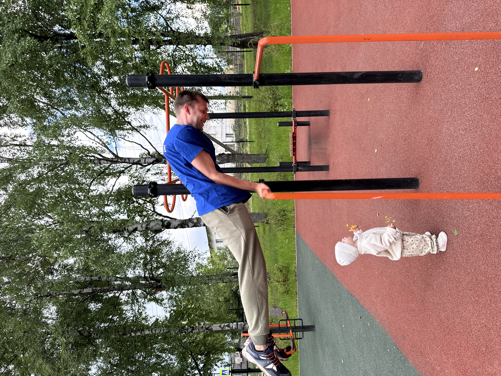 An outdoor scene depicts a man hanging from exercise bars at a park in Вассята, Russia, while a toddler with flowers stands nearby on the ground. The man is dressed in a blue shirt and gray pants and appears to be exercising. The toddler is wearing light-colored clothing. The setting includes a playground with a red and green safety surface and various exercise bars, surrounded by trees. A building is partially visible in the background.