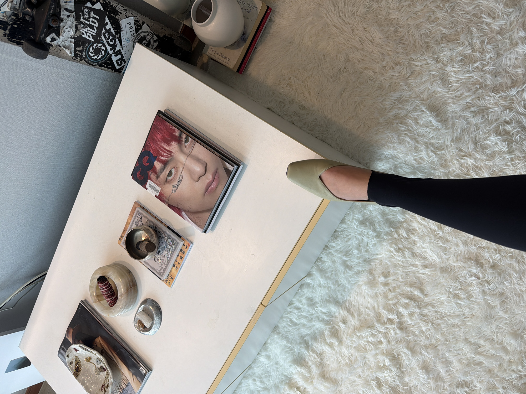 This eye-level, overhead shot captures a scene within Seoul, South Korea. A white coffee table dominates the frame, displaying magazines, a small bowl with a lit stick of incense, and other decorative items. At the top right of the coffee table sits a white vase on top of books. A leg, clad in black leggings and a light green shoe, extends onto the rug from the right side of the table. The rug, a plush, white shag, covers a portion of the floor. Adjacent to the coffee table on the left sits a gray sofa with a skateboard resting on the back. The focus remains on the objects and surfaces, without directly showing any people.