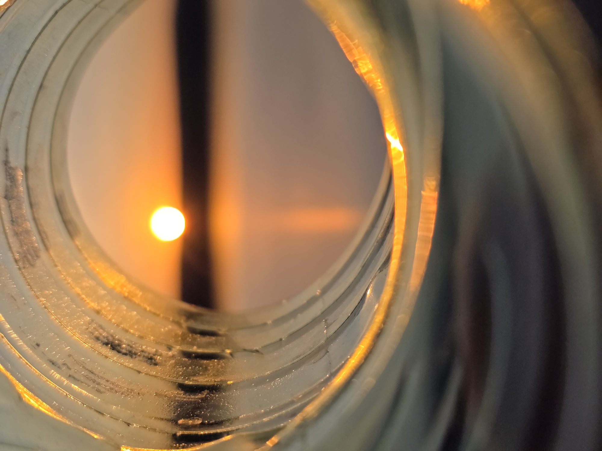 The image shows a sunset viewed through the interior of a transparent, cylindrical object, likely plastic, with multiple concentric rings. The sun is a bright, circular orb positioned centrally in the background, appearing between a dark vertical stripe. The foreground consists of the textured surface of the cylinder, with light refracting and reflecting along its edges. The overall scene is bathed in warm orange and yellow tones from the setting sun. The image's precise location in Chaikovsky, Russia cannot be determined based on the provided visual information.