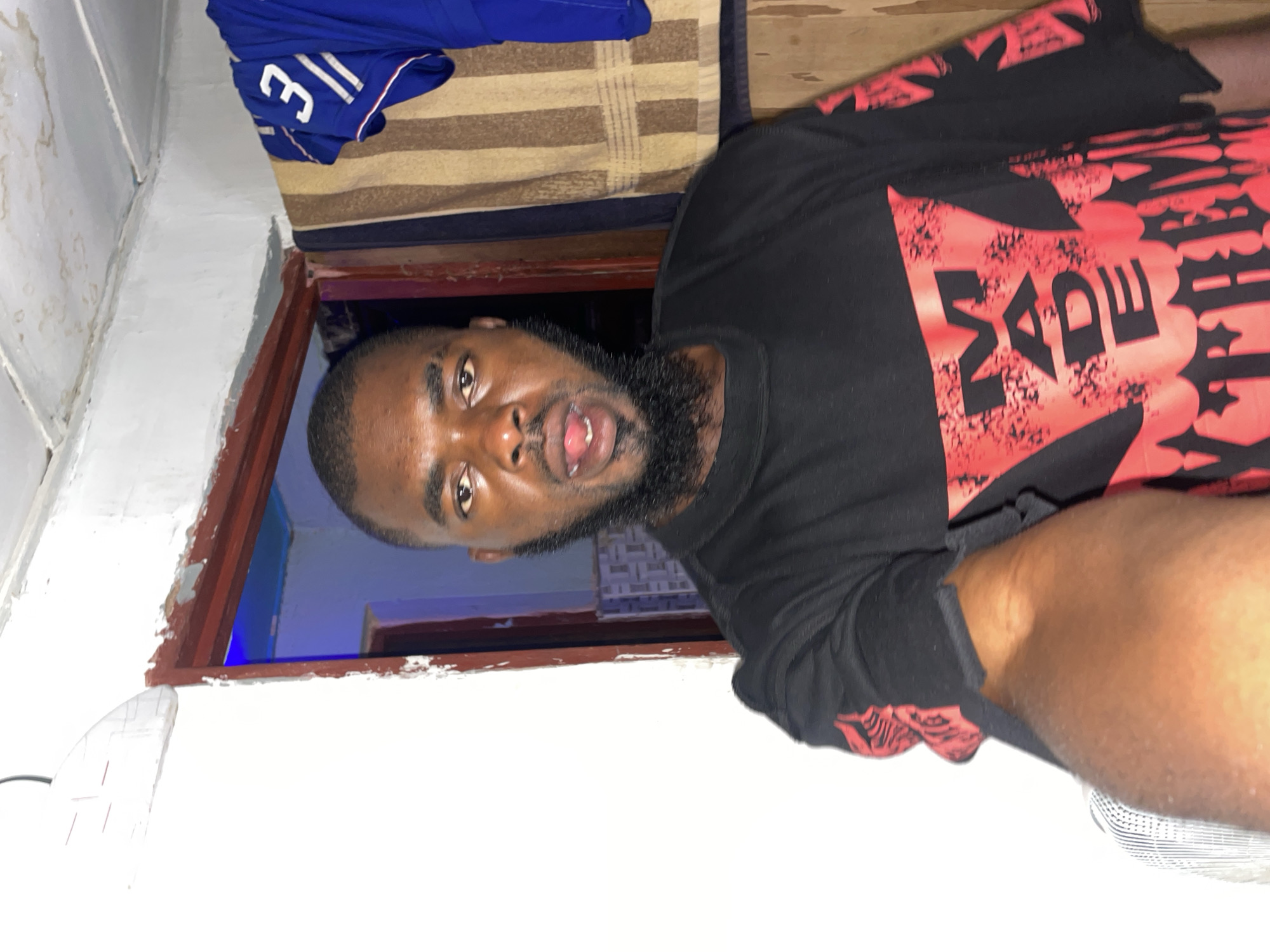 An indoor, close-up selfie captures a dark-skinned male subject, approximately in his 20s to 30s, looking directly at the camera with his mouth slightly open, revealing his tongue. He has a full beard and short hair. He is wearing a black t-shirt with a prominent red graphic design on the chest and sleeves, featuring the word "MADE" in a distressed font, likely part of a larger phrase. His right arm is partially visible in the lower left, holding the camera.
The background consists of a white wall on the left, displaying minor wear and a white, horizontally-lined object near the top. A dark wire descends from the top left corner. To the right of the subject, a doorway with a dark brown, peeling frame opens into a dimly lit room. Inside this room, a blue light source illuminates the upper wall and ceiling, and a patterned wall surface is visible. Further back within the doorway, another opening or darker room is present. To the right of the doorway, a striped fabric, possibly a towel or blanket with brown, tan, and dark stripes, hangs over a wooden or panelled wall section. A blue garment with the white number "3" and white stripes is draped over the striped fabric in the upper right. The ceiling above is white with visible seams and some discoloration. The scene is described as being in the city of Jos, Nigeria.