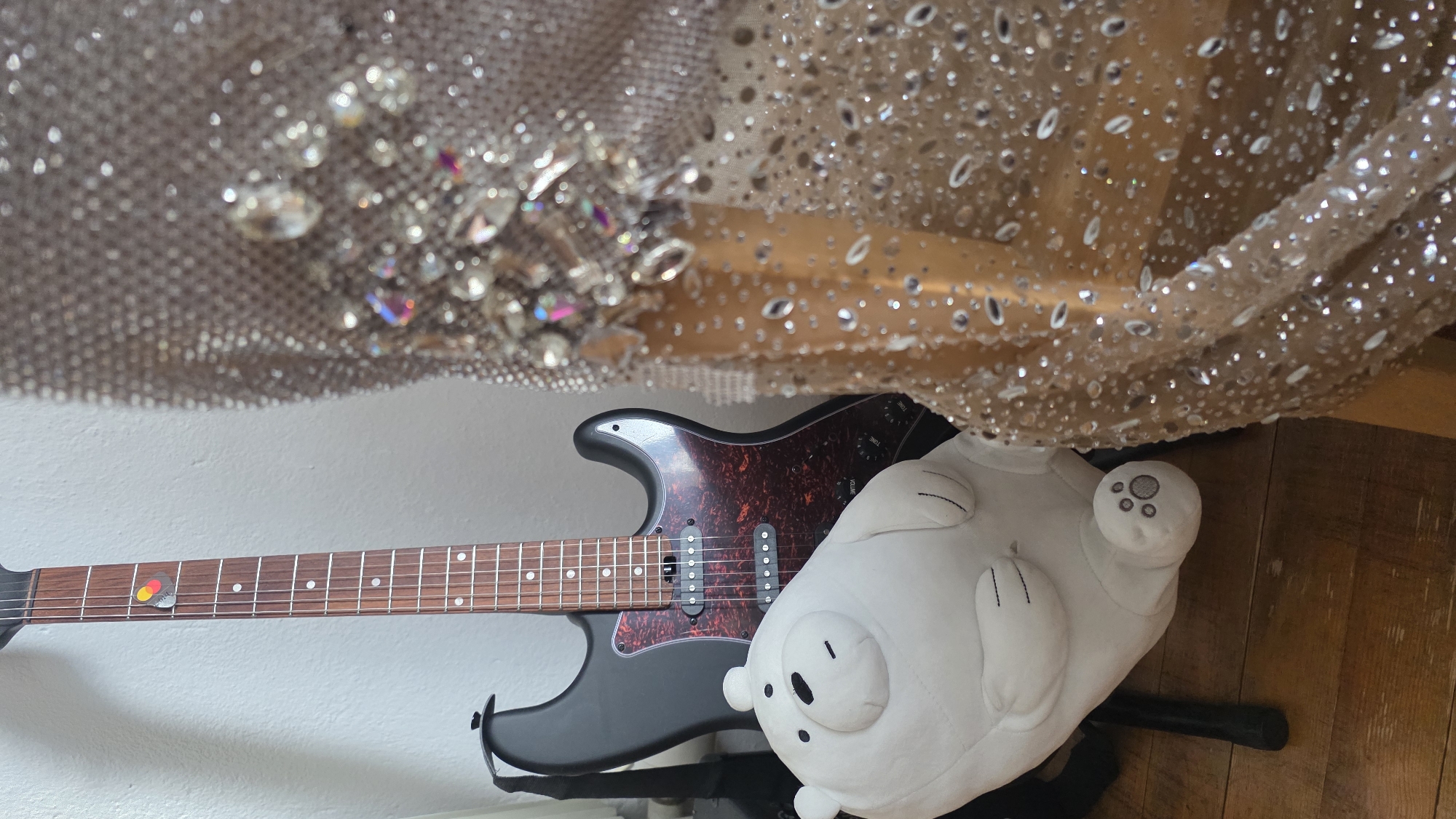 The image shows a black electric guitar resting against a white wall, next to a fluffy white plush toy. A shimmering, possibly beaded or sequined, fabric hangs overhead. The guitar has a reddish-brown fretboard, and the pickguard appears to be a marbled red color. A guitar pick is visible, resting on the neck near the headstock. The background indicates a domestic interior, with wooden flooring partially visible. There is no information available in this image to determine its location in the city of Blagoevgrad, Bulgaria.