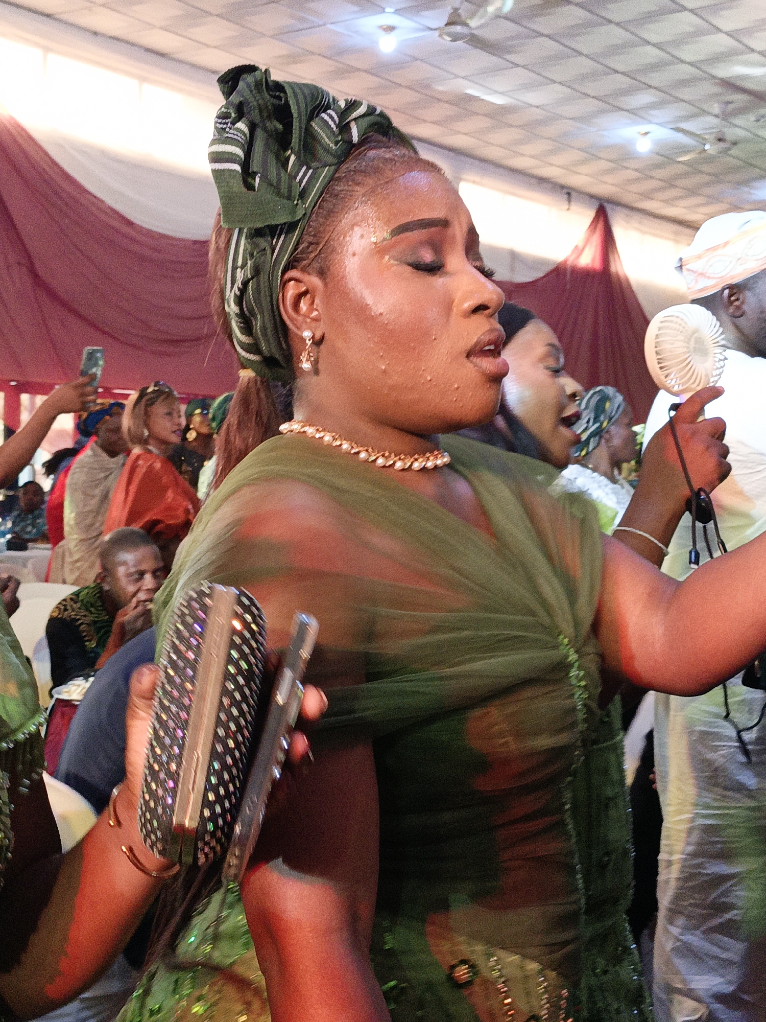 The image depicts a woman singing at an event, likely a celebration, in Fagge D2, Nigeria. She is wearing a green headwrap and a matching green dress with a sheer overlay, accessorized with a pearl necklace and dangling earrings. She holds a shimmering clutch purse and a mobile phone. The woman appears to be sweating. Behind her, another woman sings and a man holds a small fan. Other people are visible in the background, some holding up phones to record the event. The setting appears to be an indoor event space with draped fabrics in the background.