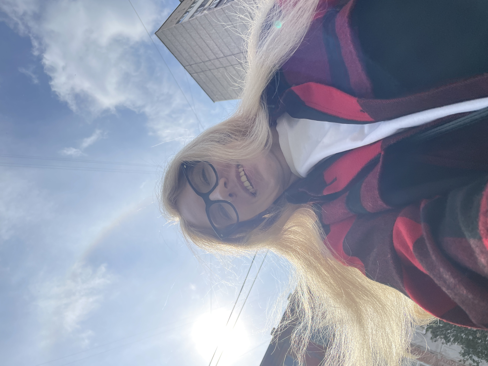 A low-angle selfie captured outdoors in Izhevsk, Russia, features a smiling person with long, bright blonde hair and black-rimmed glasses, looking down towards the camera. They are wearing a red and black plaid shirt over a white top.
The dominant background is a clear, bright blue sky with scattered white clouds, indicating daytime. A powerful sun flare emanates from the lower right portion of the frame, with the sun appearing directly overhead or slightly behind the subject. A faint, circular atmospheric phenomenon, resembling a halo or sun dog, is visible in the sky to the left of the person's head. Parts of a grey, multi-story building with a grid-like pattern are visible in the upper left and bottom right corners, along with thin overhead wires, suggesting an urban setting. The person's broad, open-mouthed smile conveys a cheerful or happy emotion. There is no visible text in the image.