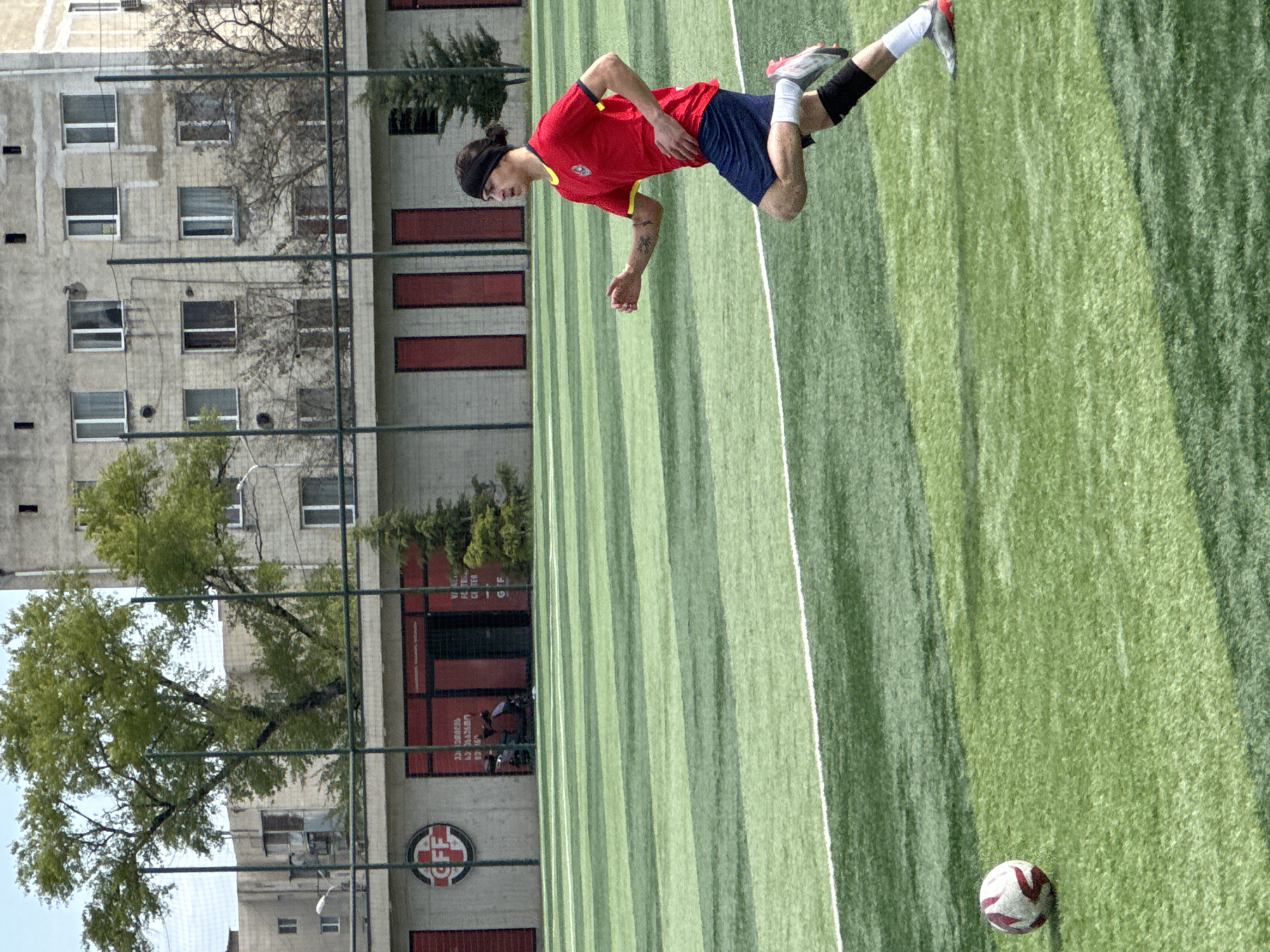A soccer player in a red shirt and blue shorts is preparing to kick a soccer ball on a green turf field. A building with multiple windows and red vertical elements is visible in the background behind a chain-link fence. Trees are also present in the background. The image appears to be taken at the Football Federation Academy in Tbilisi, Georgia, as indicated by the "CFF" logo visible on the building. The sky is clear and the lighting is bright.