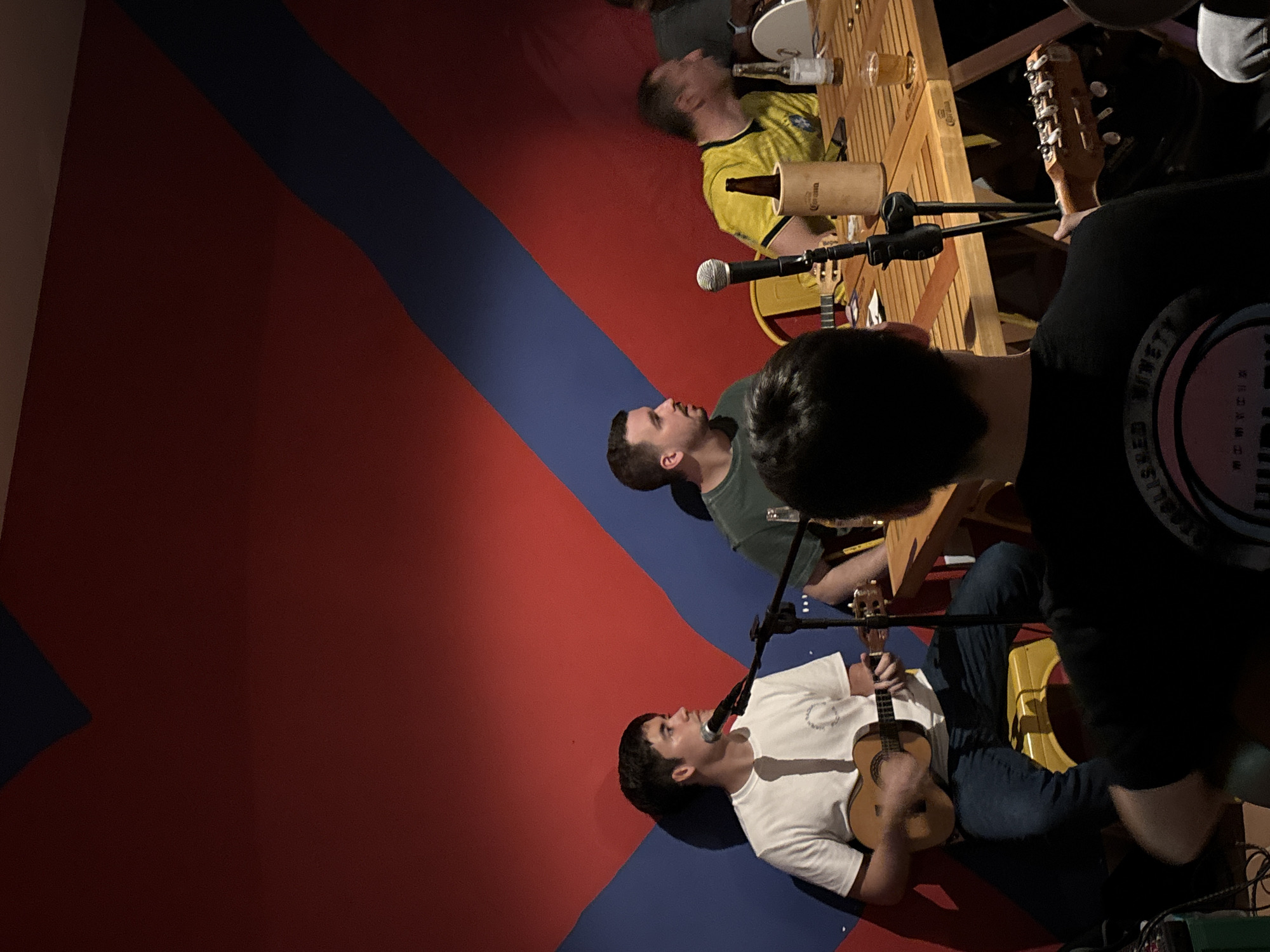 The scene shows four individuals performing at a venue. The backdrop consists of a wall painted with a large red area and a diagonal blue stripe. The venue might be in São Paulo, Brazil, but there is no element in the image that could confirm this information. Three of the individuals are seated at a wooden table, with microphones positioned in front of them. One person wears a yellow shirt, another a green shirt, and the third a white shirt and is playing a ukulele. The fourth individual stands to the right and is partially obscured, wearing a black shirt. On the table are bottles and glasses. Musical instruments and equipment are also visible.