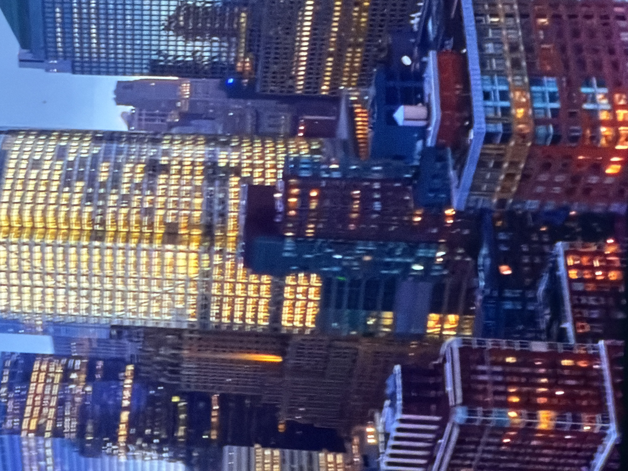 This image depicts a dense urban cityscape at dusk or night, located in the city of Maidstone, United Kingdom. The scene is dominated by numerous multi-story buildings and skyscrapers, predominantly illuminated by internal lights, against a dark blue twilight sky.
In the center-right, a tall, light blue tinted skyscraper displays a uniform grid of consistently lit windows. Adjacent to it on the center-left, a prominent, gold-hued skyscraper exhibits a bright, nearly full illumination of its many windows, casting a warm glow. In the foreground on the bottom-left, a red-brick building with multiple illuminated windows is visible, featuring a light-colored structure on its roof. To the bottom-right, another red-brick building stands, distinguished by a light blue architectural band near its upper levels and a white, cylindrical water tower positioned on its roof. Various other darker buildings are present in the midground and background, some with sparse illumination, including one slender building in the center with a vertical orange light emanating from its upper section. No individuals, specific actions, or interactions are discernible within the frame.