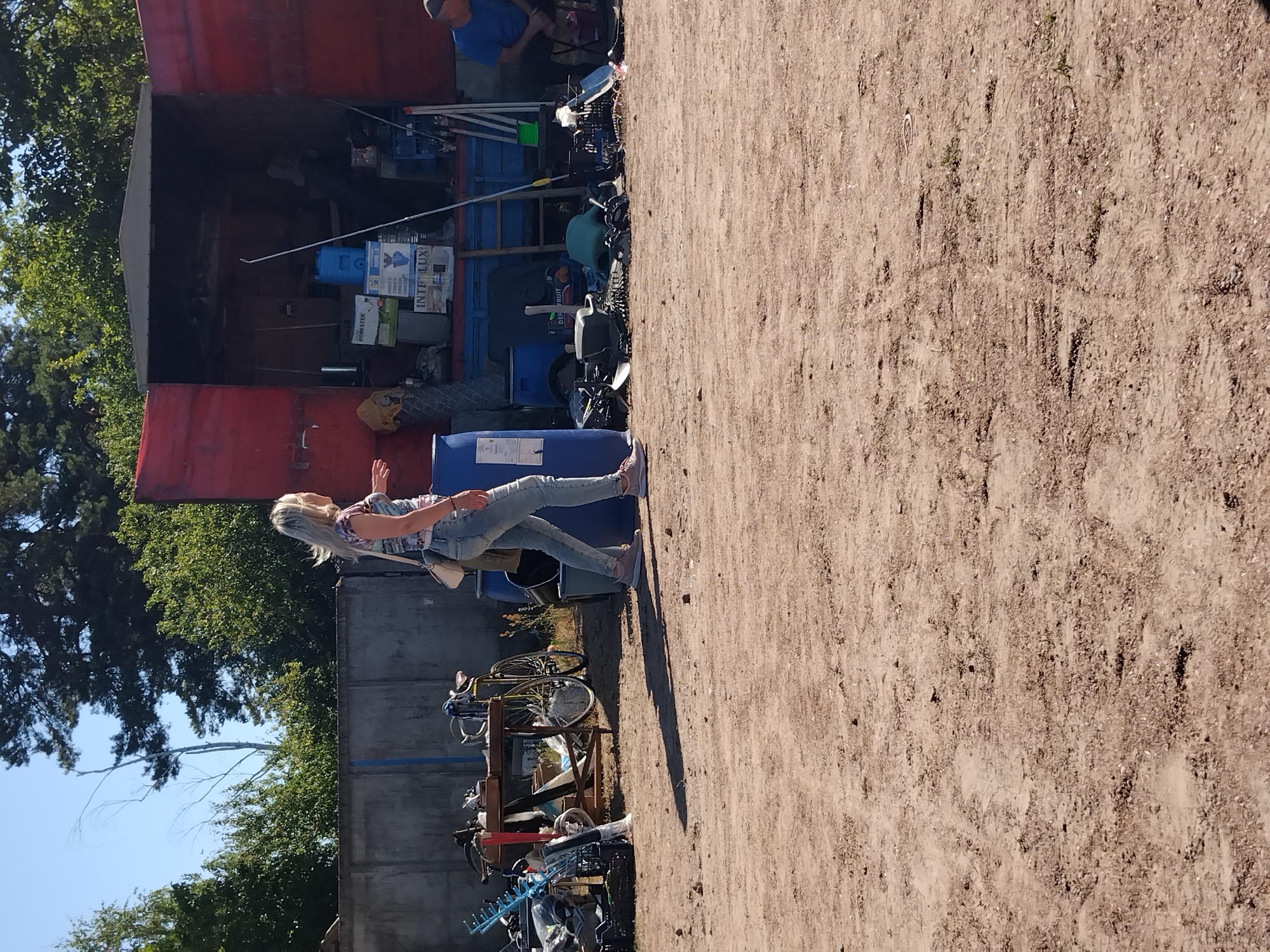 The image depicts an outdoor scene in Irshansk, Ukraine, likely a junkyard or a makeshift marketplace. In the foreground, a woman with blonde hair, wearing a patterned top and light-wash jeans, walks away from the camera towards a red trailer. She has a white crossbody bag. The ground is composed of dirt and gravel. Behind her, various items are strewn about, including a blue plastic barrel, a bicycle on a stand, and piles of scrap metal and other miscellaneous objects. To the left, a concrete wall is visible, with trees and foliage beyond it. The red trailer, open at the back, is filled with boxes, containers, and more assorted items. A person in a blue shirt sits in the right background, facing away from the camera. The sky above is clear and blue, suggesting a sunny day.