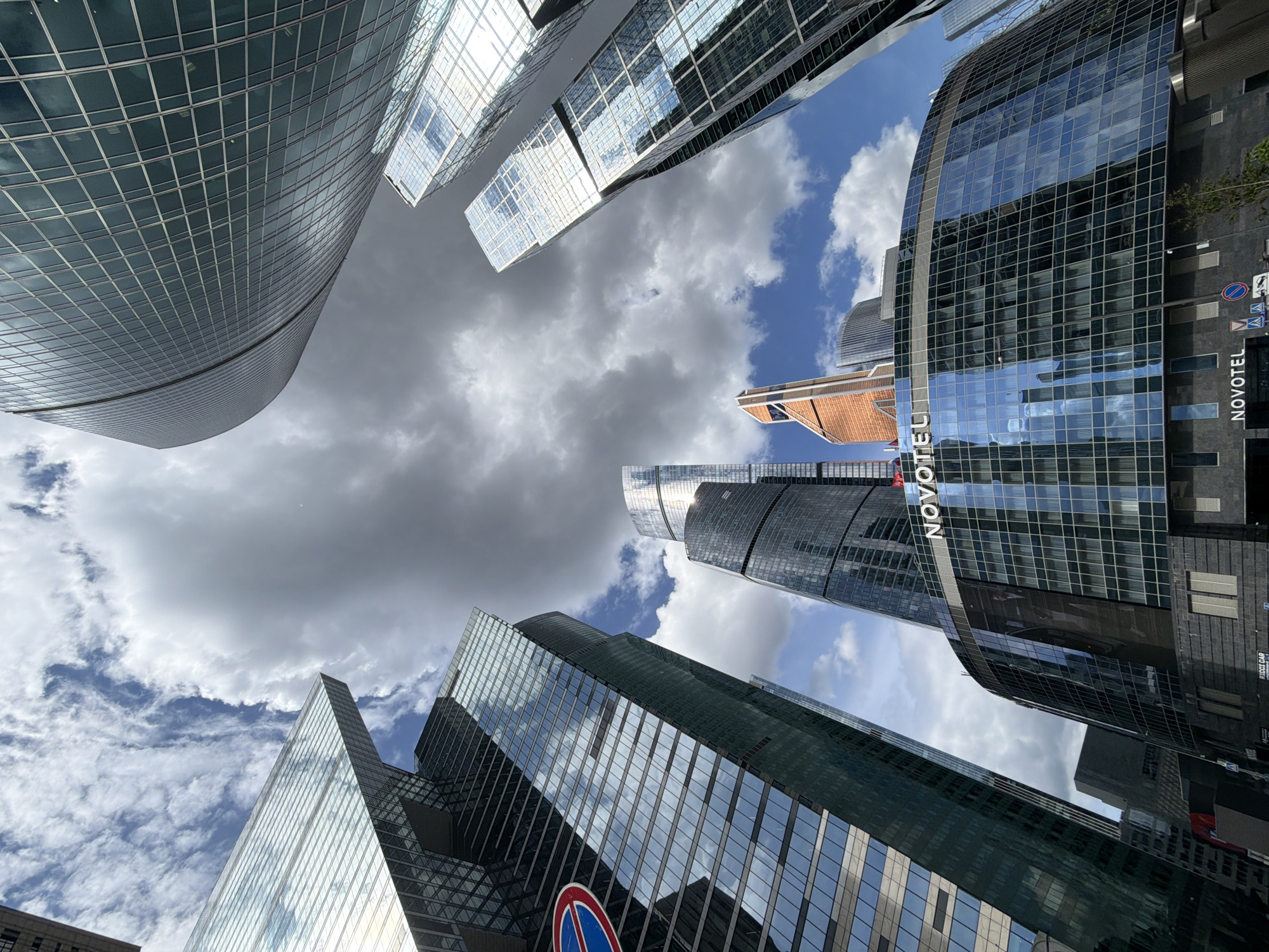 The image is a low-angle shot of several skyscrapers in Moscow, Russia. The architecture is modern and features glass curtain walls reflecting the sky. A building with the name "NOVOTEL" is visible. The sky is partially cloudy, with patches of blue sky visible. No people are visible in the scene.