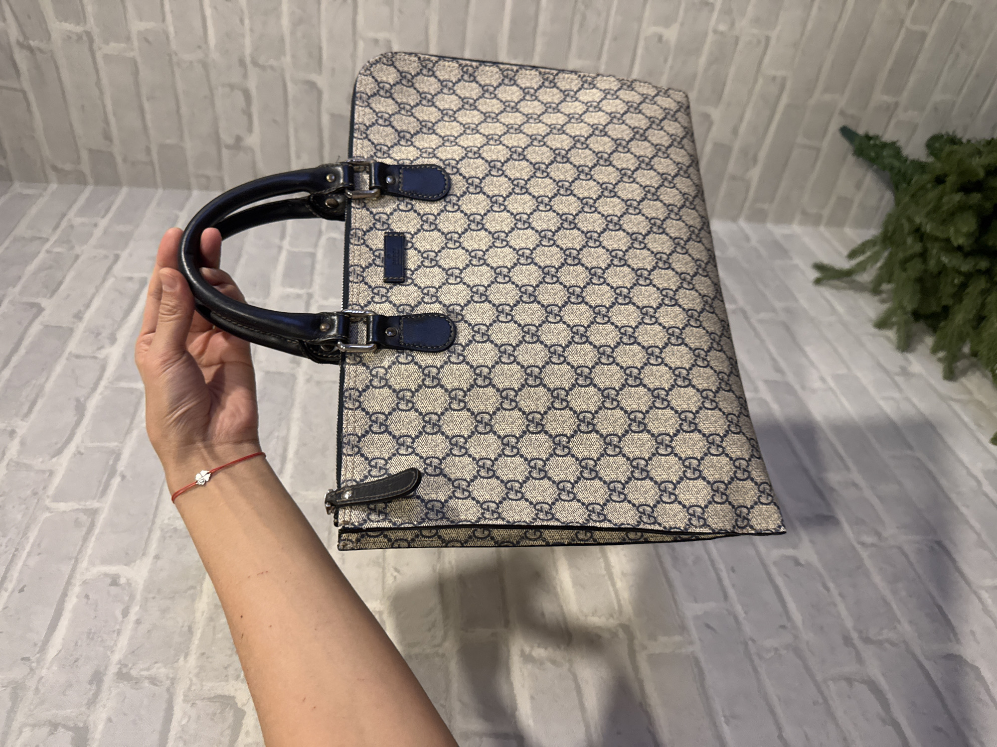 An eye-level indoor shot in Bangkok, Thailand, features a person's arm and hand holding a beige and navy blue Gucci handbag. The handbag, held upright, displays a repeating GG monogram canvas pattern. It has dark blue leather handles and trim, accented with silver-toned metal hardware, including small buckles attaching the handles. A small rectangular leather tag on the front of the bag, below the top handle, bears the faint inscription "GUCCI made in Italy."
The person's forearm and hand are visible, with a thin red string bracelet adorned with a small, silver-toned, four-leaf clover charm on the wrist. The background consists of a light-colored, possibly white or light gray, brick-patterned wall. To the right, a blurred section of green artificial foliage, resembling a decorative plant or part of a Christmas tree, is partially visible. The lighting appears even, suggesting an indoor setting with artificial light, designed to highlight the handbag. The image's focus is on showcasing the details of the luxury accessory.