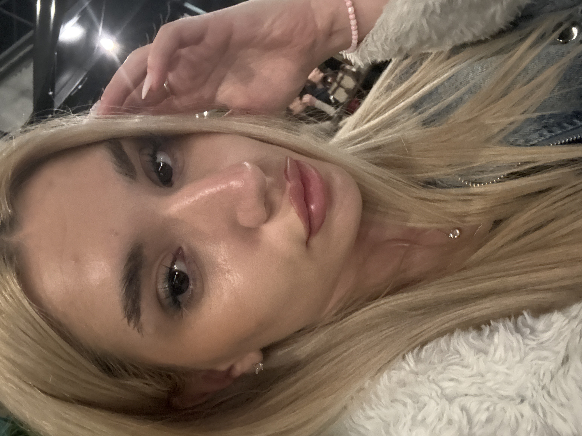 A close-up selfie shows a young woman with long, flowing blonde hair, light skin, and a neutral, serene expression. Her head is tilted, and she gazes directly into the camera with her eyes open and lips slightly parted, accentuated by lip gloss. Her eyebrows are well-defined, and she wears mascara and eyeliner. One hand, adorned with manicured nails (light-colored, almond shape), is gently placed near her forehead, touching her hair.
She is wearing a light blue denim jacket featuring a prominent white, fluffy faux-fur collar or lining, which is visible around her neck and shoulder. A delicate silver-toned necklace with a small, sparkly pendant is visible on her neck, along with a small stud earring in her left ear. On her right wrist, she wears a light pink beaded bracelet.
The setting appears to be indoors, likely in the evening or at night, indicated by the dark, out-of-focus background with bright, diffused artificial light sources. The lighting creates highlights on her skin, particularly on her forehead, nose, and lips. The activity depicted is a self-portrait, possibly taken while resting or posing casually. There is no visible text in the image. The photo was taken in Sofia, Bulgaria.