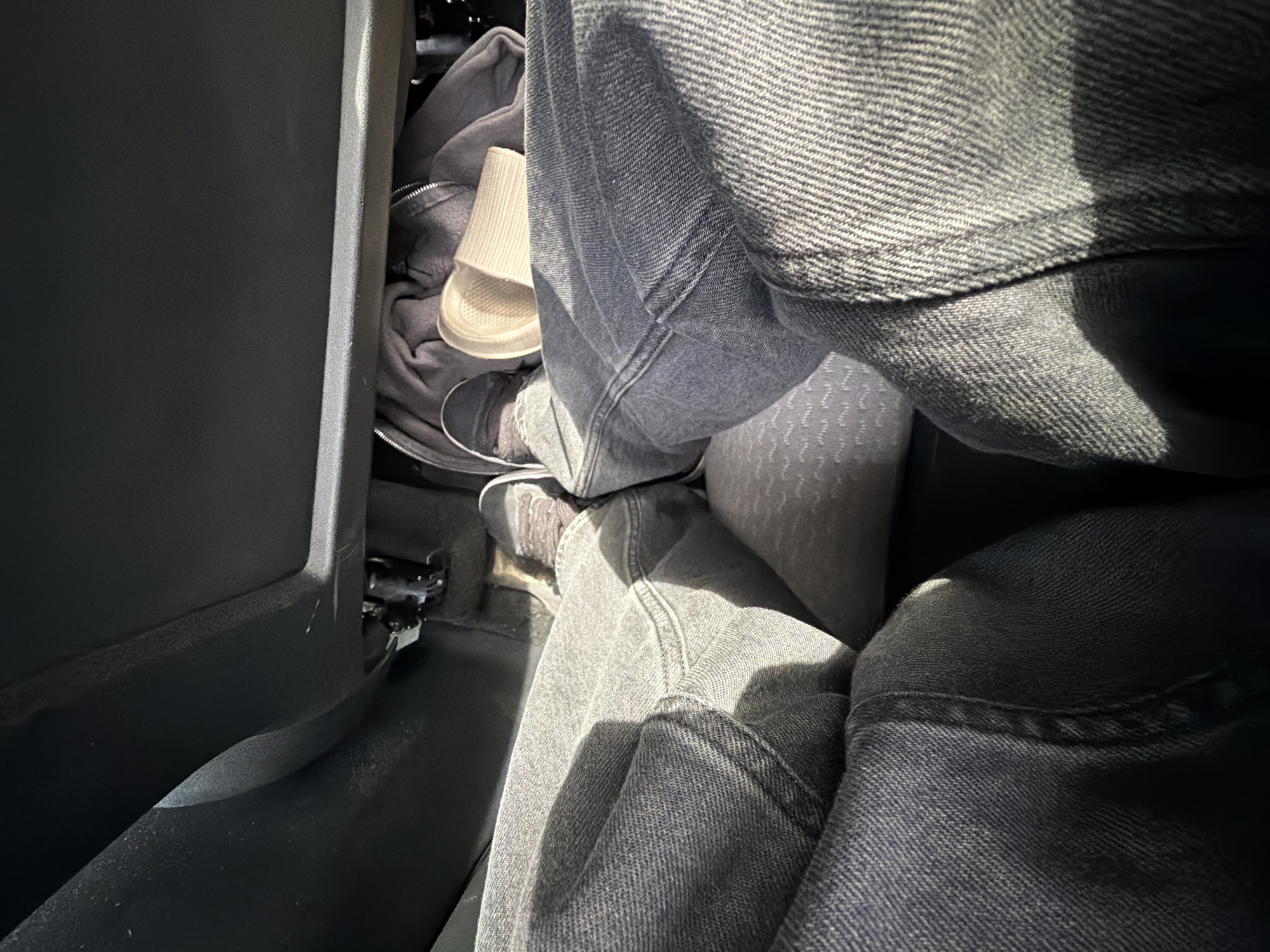 The image depicts a close-up, overhead view of a person's legs and feet inside what appears to be the passenger footwell of a vehicle. The person is wearing dark wash denim jeans and grey sneakers. Their legs are crossed, with one foot resting on the floor and the other slightly elevated, possibly on the seat or the floor mat. Beneath the feet, a grey fabric object, possibly a jacket or blanket, is visible, along with a light-colored, ribbed object that could be a paper cup or a similar disposable container. The interior of the vehicle is dimly lit, with strong sunlight casting shadows across the jeans and the floor. There is no indication of the location being in Vallensbæk Landsby, Denmark.