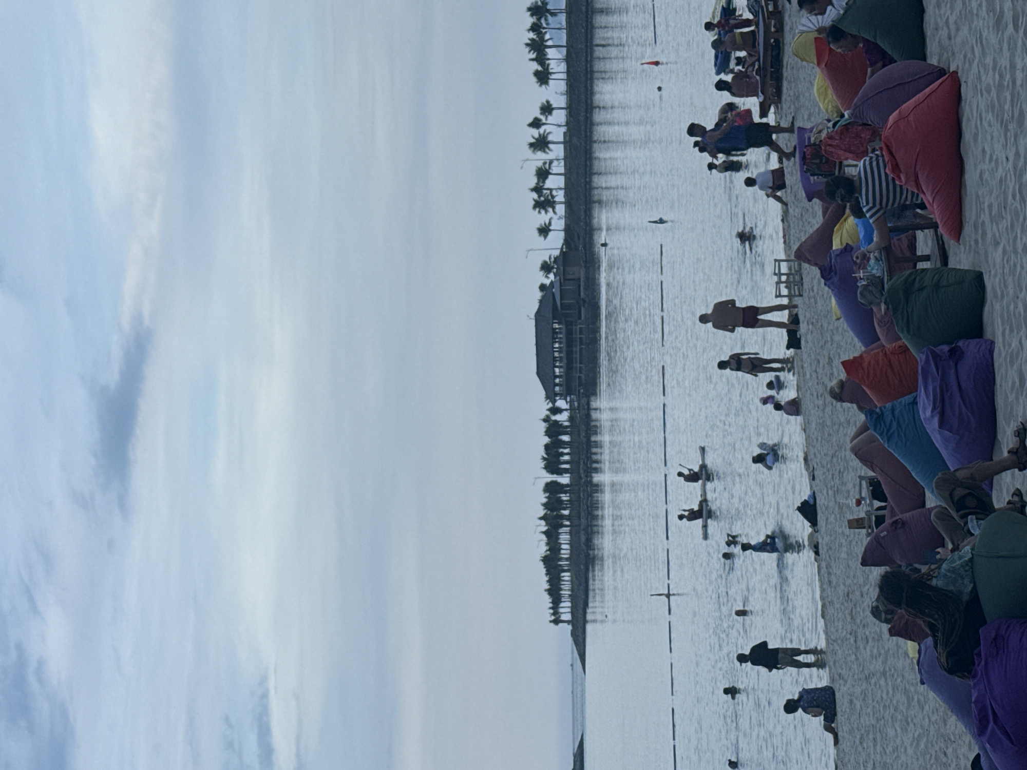 The image depicts a beach scene, likely in the city of Bac Nha Trang Ward, Vietnam. The sky is overcast. The body of water is calm. People are wading, swimming, and kayaking in the water. Colorful beanbag chairs are scattered along the sandy beach. Some people are sitting and relaxing on the beanbag chairs. Palm trees line the waterfront in the distance.