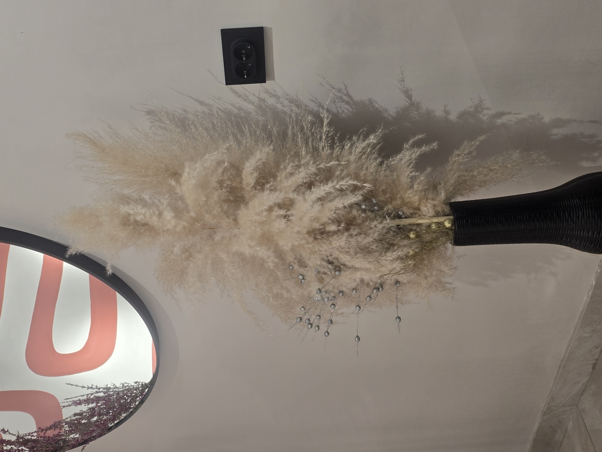 The image shows a tall, light-colored dried grass arrangement in a black vase, placed against a plain, light-colored wall. To the left, a portion of a circular mirror with a coral-colored abstract design is visible, with some dried purple flowers cascading over its edge. To the right of the grass arrangement, a black electrical outlet with two sockets is mounted on the wall. The lighting creates a shadow of the grass on the wall to the right. The floor is visible at the bottom, showing a light-colored tiled surface and a darker baseboard. This scene is indoors, likely in a residential or commercial space, and no specific location in Ulan-Ude, Russia can be determined from the image. There are no people or animals present.
