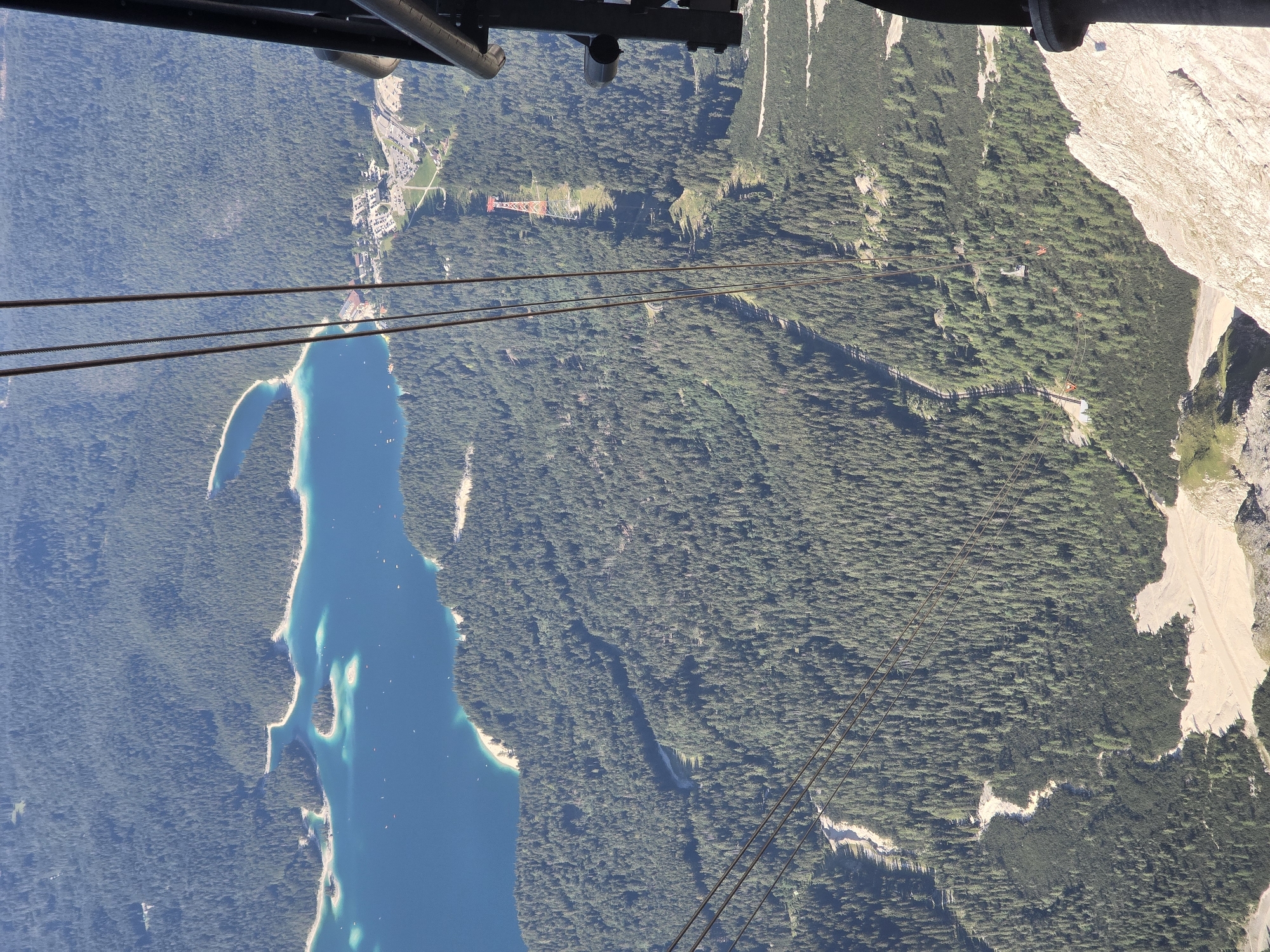 The image captures a high-altitude view of the Eibsee lake and surrounding forest near Garmisch-Partenkirchen, Germany, as seen from a cable car. The turquoise waters of the Eibsee are visible in the upper left quadrant, dotted with small boats. The lake is fringed by a lush green forest that covers the mountainous terrain. In the mid-ground, a red and white cable car tower stands prominently, indicating the path of a cable car system. Further down the slope, a cluster of buildings and a parking lot are visible, suggesting a tourist area. Several cable cars, some with orange cabins, can be seen suspended on cables, traversing the landscape. The foreground consists of a rocky, mountainous outcrop, characteristic of alpine environments. The overall scene depicts a natural landscape enhanced by recreational infrastructure.