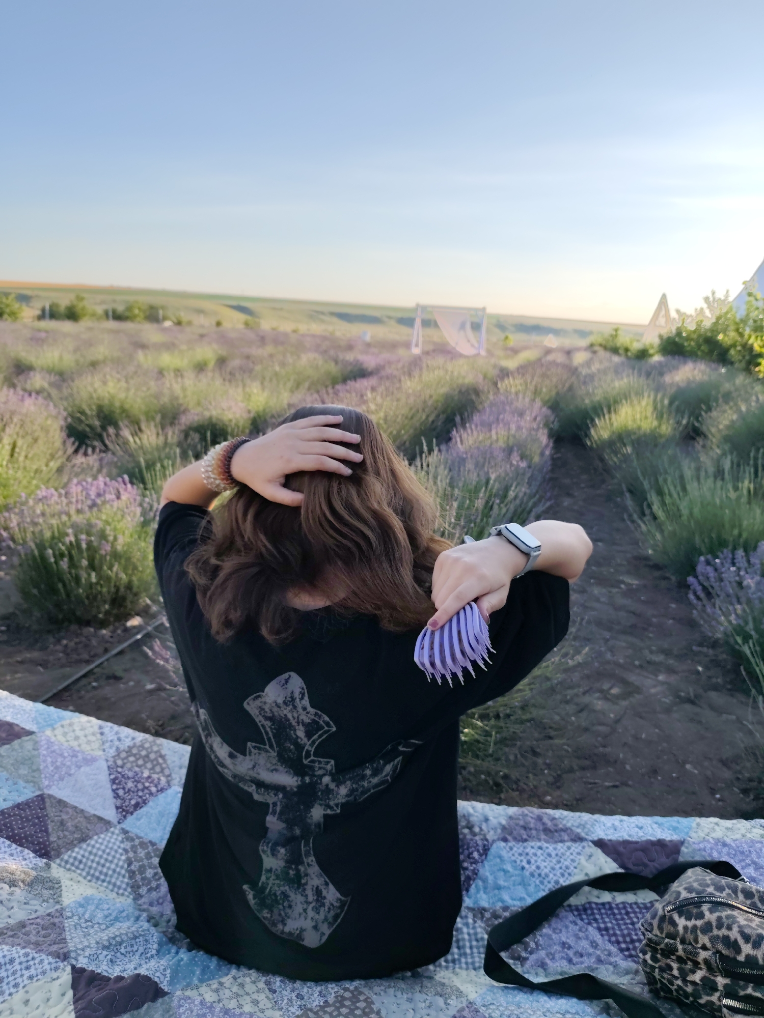 A person sits on a quilt in a lavender field, brushing their hair. The person is wearing a black t-shirt with a large, faded graphic design on the back. They are also wearing a watch on their left wrist and several beaded bracelets on their right arm. The person is seated on a colorful patchwork quilt, and a leopard-print bag with a black strap rests beside them.
The setting is a vast lavender field, with rows of purple lavender stretching into the distance under a clear blue sky. In the background, there are several white tent-like structures and some trees. The sun appears to be low in the sky, suggesting either early morning or late afternoon, casting a warm light over the scene. The overall atmosphere is peaceful and serene.
The image was taken in Nechaiane, Ukraine.