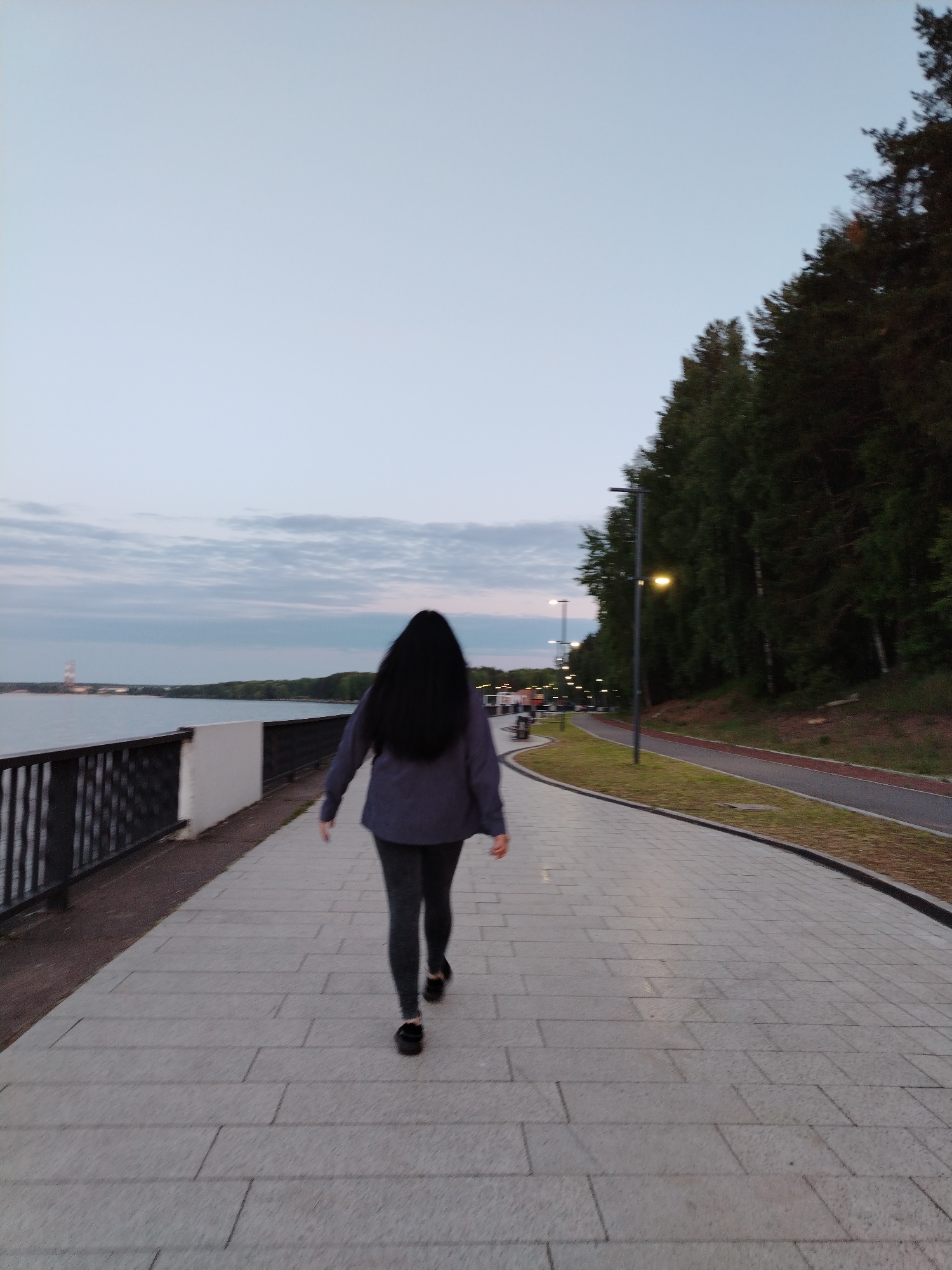 The image depicts an evening scene on a paved promenade in the city of Chaikovsky, Russia. A solitary individual, viewed from behind, is walking away from the camera. This person has long, dark hair and is dressed in a dark jacket and dark leggings, with black footwear. The promenade, constructed from light grey rectangular paving slabs, extends into the distance. To the left, a dark metal railing borders a vast, placid body of water, likely the Votkinsk Reservoir, which reflects the pale, cloudy sky. Across the water, a distant tree-lined landmass is visible, with a tall, light-colored structure, possibly industrial or a tower, prominent on the far left horizon. To the right of the promenade, a narrow strip of grass precedes a secondary, darker paved pathway, featuring a distinct reddish lane marking, indicating a dedicated track. Beyond this pathway, a dense line of tall, dark green trees forms a forest, with scattered lighter-barked trees visible within the foliage. Multiple streetlights with illuminated yellow lamps are positioned along the pathways, providing artificial illumination as dusk settles. The sky is a soft gradient of light blue and grey, characterized by diffuse cloud cover.