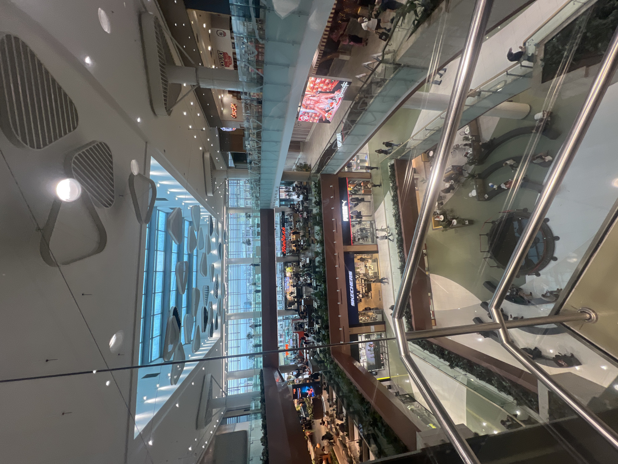 The image depicts the interior of a multi-story shopping mall. The mall features multiple levels with various shops and restaurants, and the levels are connected by escalators. Numerous people are visible on different levels, some walking, others browsing shops, and some dining. A variety of retail brands are visible, including Skechers and Puma. The mall has a contemporary design, with large windows allowing natural light to illuminate the space. There are decorative elements on the ceiling. The reflections on glass surfaces suggest that the photograph was taken from an elevated position within the mall. Based on the information available, it is not possible to identify the exact location in Sofia, Bulgaria.