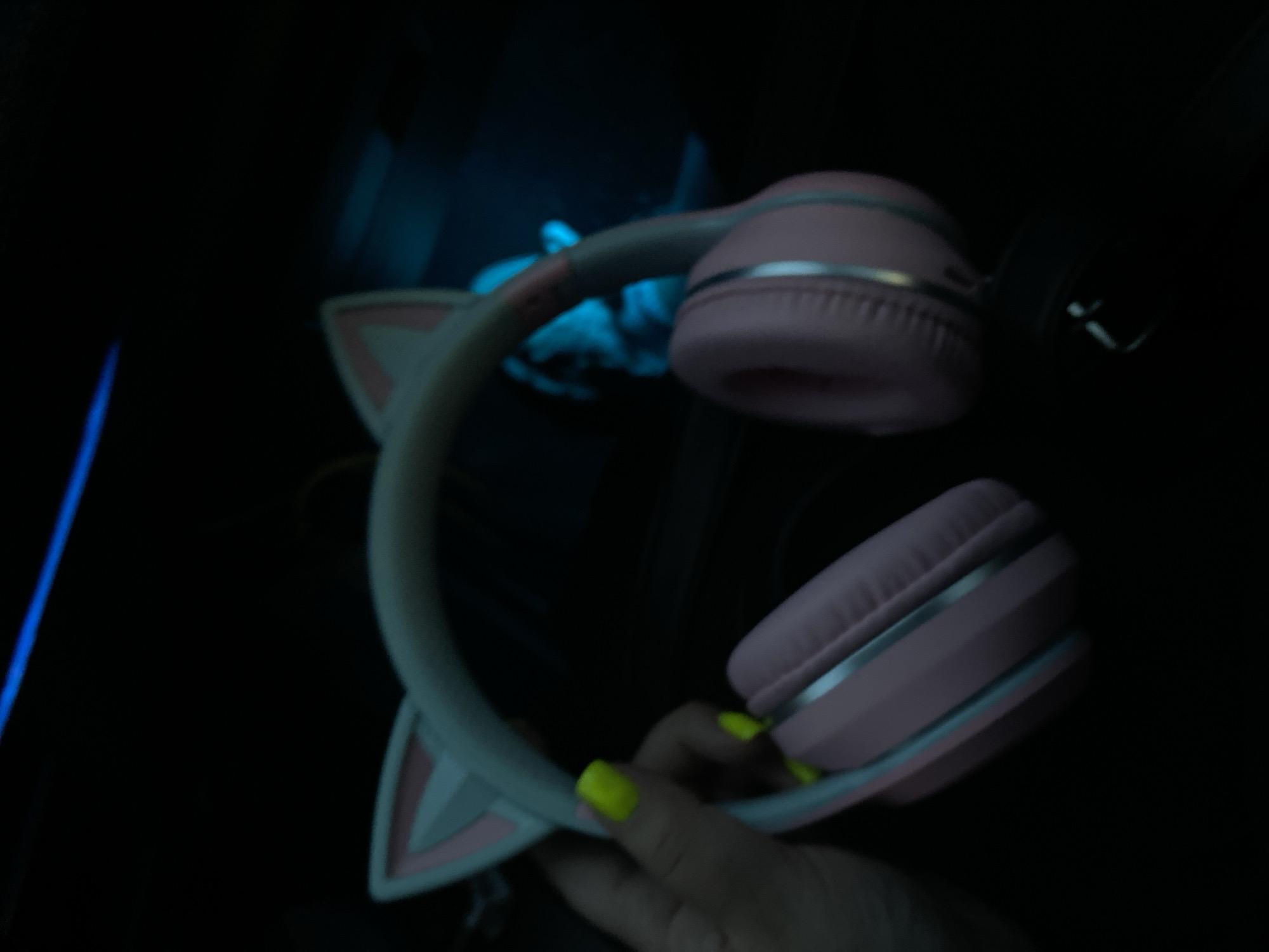 A pair of pink cat-ear headphones with light blue accents is held by a hand with bright neon yellow nail polish. The headphones are a pastel pink with light blue trim around the ear cups and along the headband. The "cat ears" on top of the headband are also pink and light blue. The background is dark, with a blurred blue light source visible in the upper right. The scene appears to be indoors, possibly in a vehicle, given the slight blur and the presence of a dark interior. The image does not provide information about the location in the city of Balashikha, Russia.