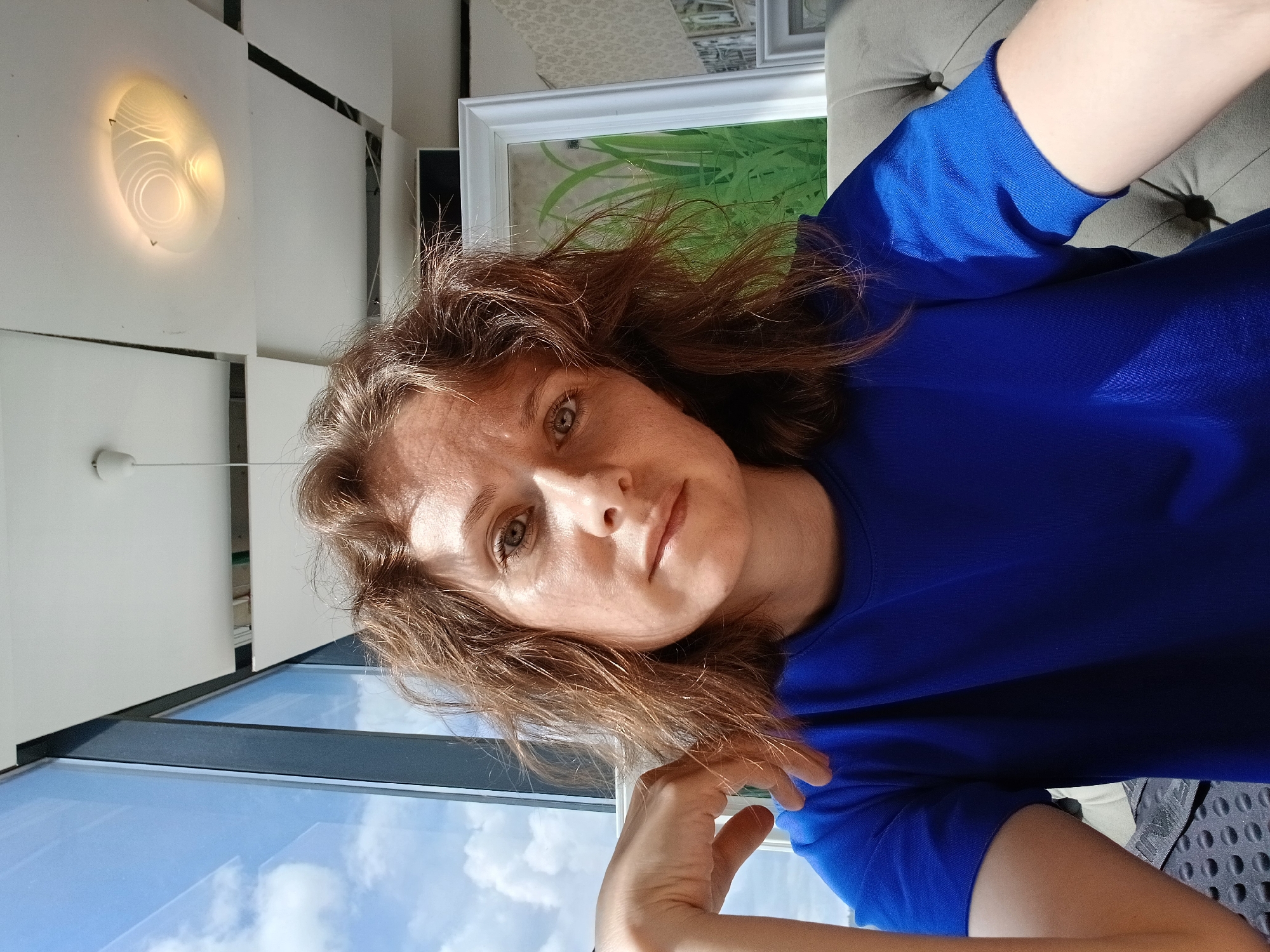 In Yekaterinburg, Russia, a woman with shoulder-length, wavy brown hair and light skin with freckles is taking a selfie indoors. She is wearing a bright blue t-shirt and is seated on a tufted gray sofa. Her left arm is bent, with her hand resting near her chin. The background features a large window with a view of a blue sky with white clouds, a modern ceiling with square panels and a circular light fixture, and a framed picture of green grass. The lighting is natural, coming from the window, and there are some shadows visible. The overall impression is a casual indoor setting with a city view.