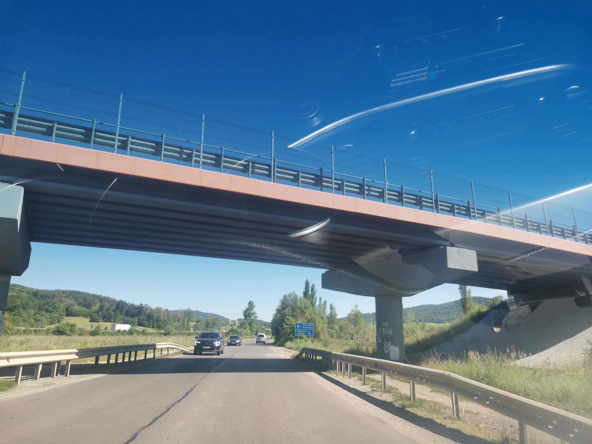 The image shows a view from inside a vehicle traveling on a highway under a bridge. The sky is clear and blue. The bridge has a brown and grey structure with railings on top. In the distance, there is a landscape of green hills and trees. A black car is visible on the highway in front, with other vehicles further ahead. Roadside barriers line the highway. Reflections are visible on the windshield of the vehicle. The blue road sign in front indicates directions to Mirovo and Radomirtsi, Bulgaria.