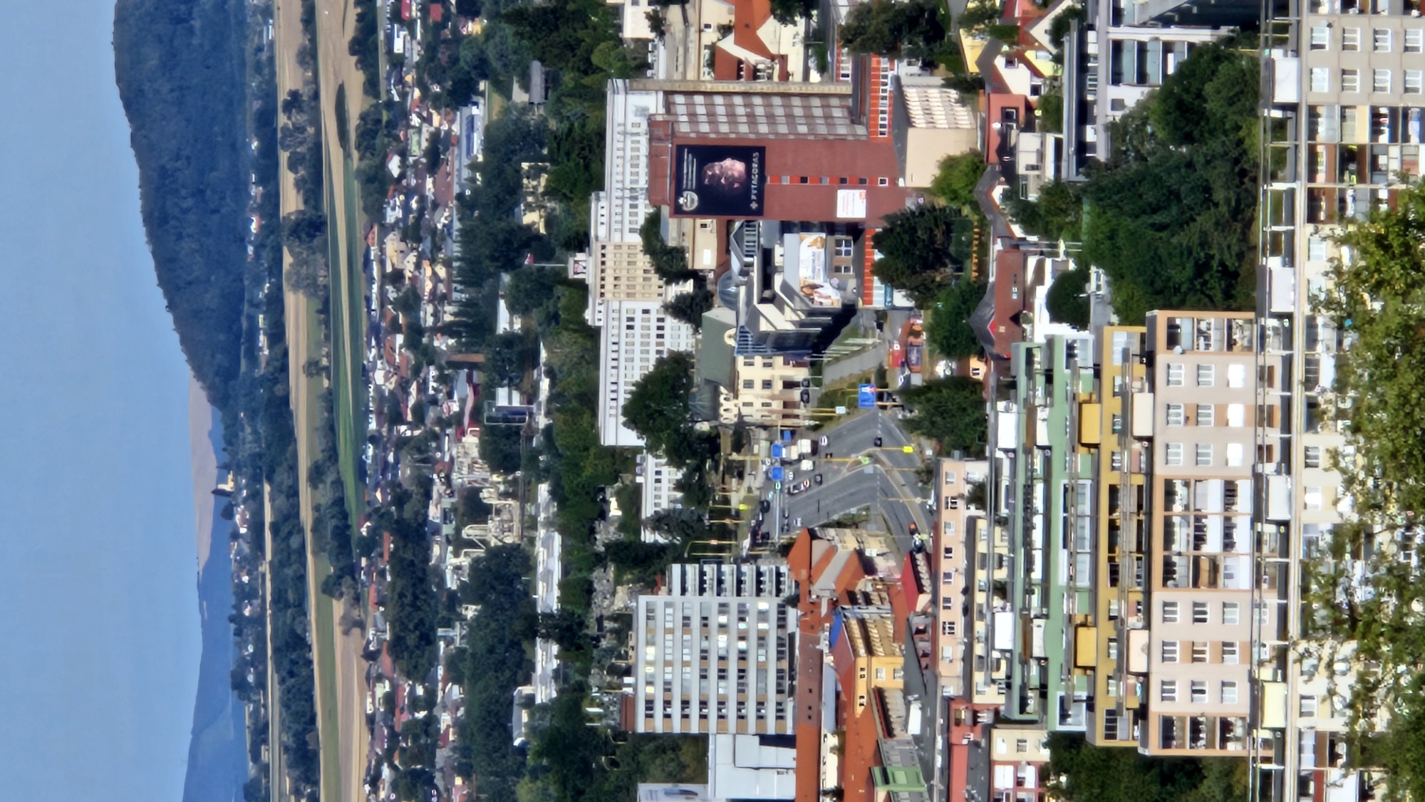 The image captures an aerial view of a densely populated urban area in Prešov, Slovakia, under a clear, bright sky. The foreground is dominated by a cluster of residential buildings, primarily multi-story apartment blocks. These buildings exhibit varied architectural styles and colors, with many featuring balconies and prominent windows. Some of these structures are painted in light hues of beige, white, and pastel colors, while others are accented with green and yellow. The apartment blocks are layered, with some appearing to be modern constructions, while others suggest an older, possibly socialist-era design, characterized by their repetitive facades and utilitarian appearance.
Further into the mid-ground, a main road cuts through the city, lined with a mixture of commercial and residential buildings. Vehicles, including cars and buses, are visible on the road, indicating active traffic. Prominent among these buildings is a tall, rectangular, red-brick building with a large advertisement poster on its side. Adjacent to it are several other mid-rise buildings, some with pitched roofs and others with flat roofs, showcasing a blend of architectural designs. One notable building in this section appears to be a church or a historical structure with a dome.
The background reveals a sprawling cityscape extending towards a gently sloping hill covered with lush green trees. Beyond the immediate urban landscape, agricultural fields and scattered houses can be seen, creating a transition from the city to the more rural outskirts. In the far distance, undulating hills and a faint outline of distant mountains under a pale blue sky complete the panorama. The overall impression is that of a lived-in city with diverse architectural elements, a functioning transportation network, and a clear demarcation between urban development and the natural landscape.