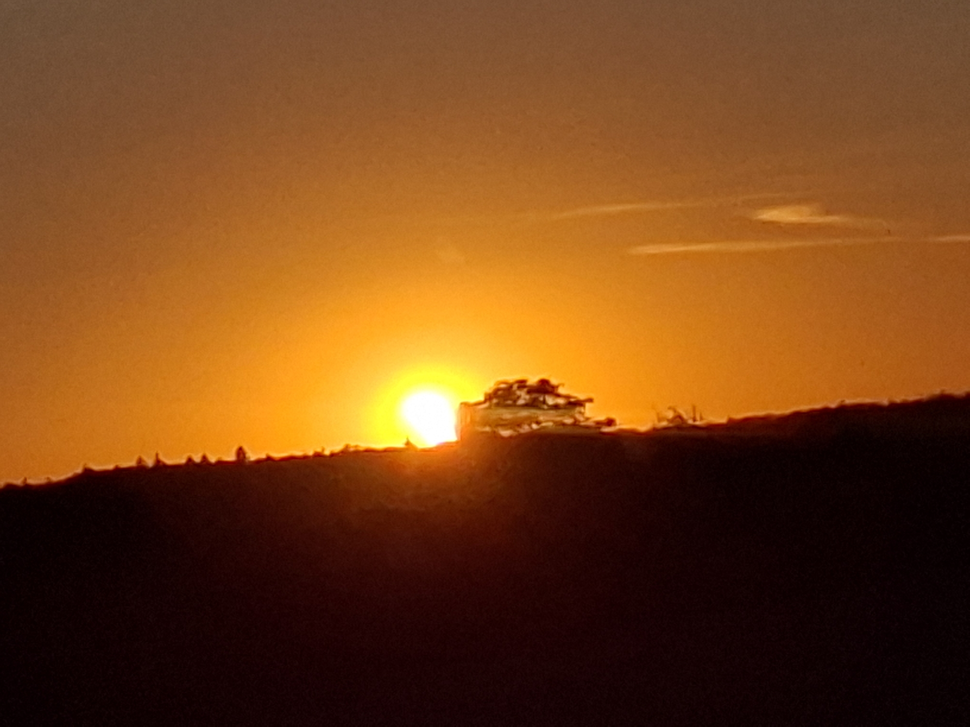A picturesque sunset is captured over the city of Sinsheim, Germany. The sun, a brilliant orb of orange and yellow, is partially obscured by the silhouette of a tree-lined ridge, casting a warm glow across the sky. Wispy clouds drift lazily across the horizon, adding depth to the scene. The foreground is dominated by the dark silhouette of the ridge, its uneven crest dotted with trees. The warm hues of the sunset create a serene and tranquil atmosphere, inviting viewers to appreciate the natural beauty of the landscape.