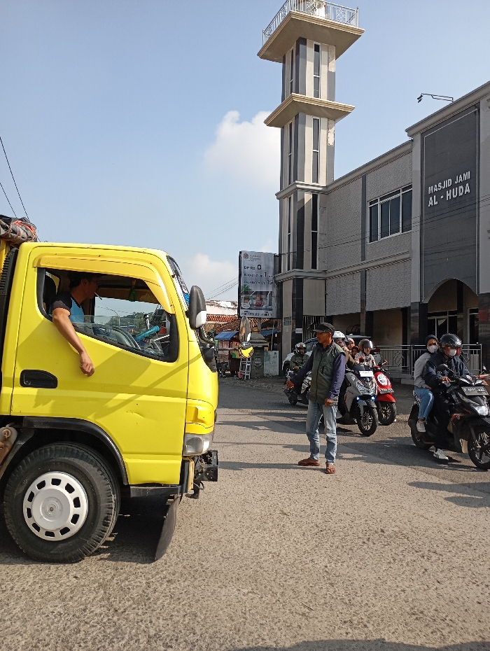 The image captures a daytime street scene in Cipatik, Indonesia. A bright yellow light-duty truck, with a male driver whose left arm rests out the open window, occupies the left foreground. Bundled items, covered by a dark tarp, are secured to the truck's cabin roof. In the midground, a male individual wearing a dark cap, an olive vest, and blue jeans stands on the asphalt road, facing left. Behind him, a queue of at least five motorcycles and scooters is visible, with riders, some wearing helmets and face masks; a dark-colored scooter with a masked rider and passenger is prominent on the far right. The background features a large, modern grey and white building labeled "MASJID JAMI AL-HUDA," flanked by a tall, multi-tiered minaret. A billboard and other structures are discernible further in the distance. The sky is clear blue with minimal cloud cover.