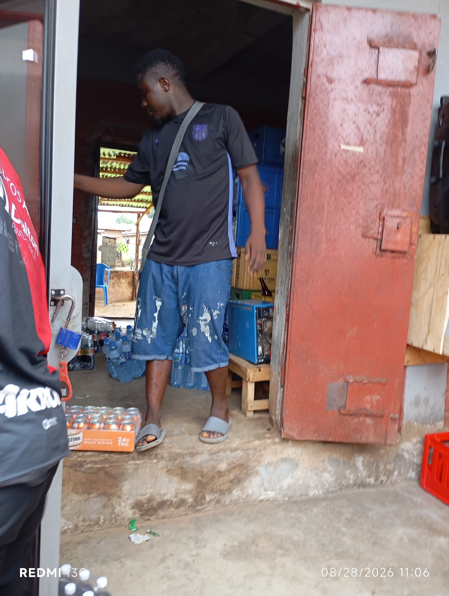 In the city of Shendam, Nigeria, a Black man is seen standing in the doorway of a structure with a large, weathered red door on the right side. He is wearing a black t-shirt with a blue emblem and denim shorts. A grey strap is slung across his shoulder. Scattered around the entrance are numerous plastic bottles of water, some stacked, and a box of canned malt drinks. To the left, another person is partially visible, wearing a black shirt with red accents. The environment appears to be outdoors, with a concrete floor and a hint of a rural or developing urban setting visible through the doorway, including some buildings and vegetation in the background. A blue plastic chair is also visible inside. The scene is lit by daylight.