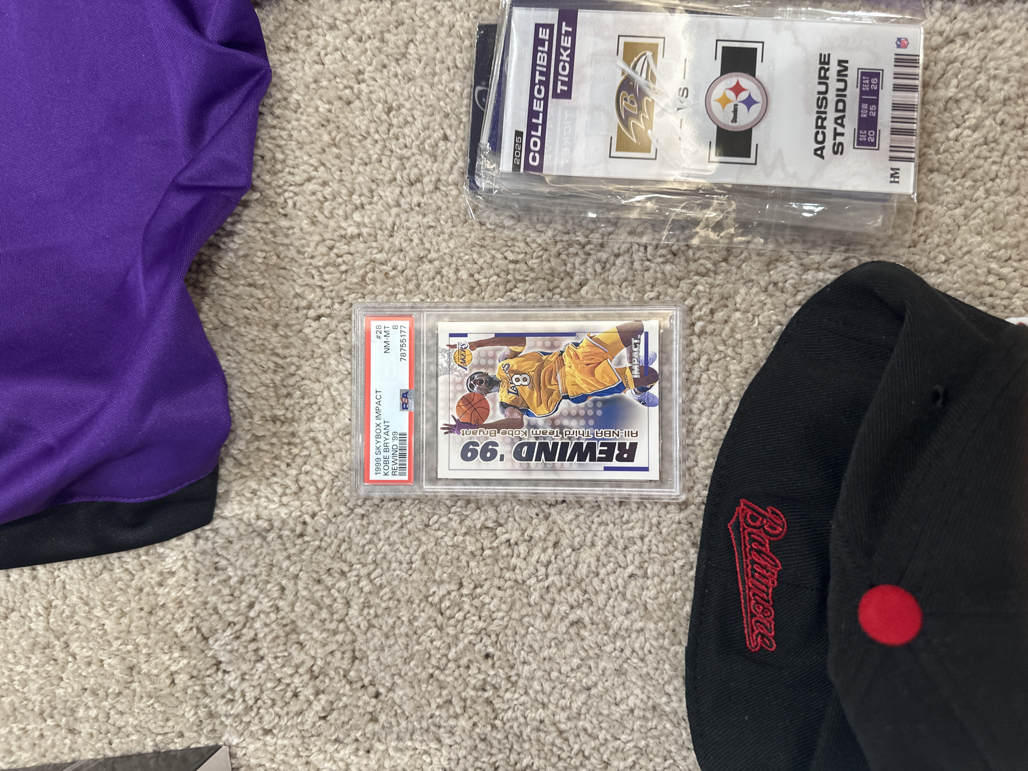 The image shows several items arranged on a beige carpet, including a purple and black garment, collectible tickets, a graded basketball card, and a baseball cap.
The collectible tickets appear to be for a Baltimore Ravens football game against the Pittsburgh Steelers at Acrisure Stadium, Row 25, Seat 26 in Section 20. The tickets are still sealed in plastic. The baseball cap is black with red accents, and reads "Baltimore" in red script. The purple garment is visible in the upper left corner. The basketball card is a graded 1999 Skybox Impact Kobe Bryant Rewind '99 card, graded NM-MT 8. The grading number is 78755177.
There are no explicit signs of Aventura, Florida.