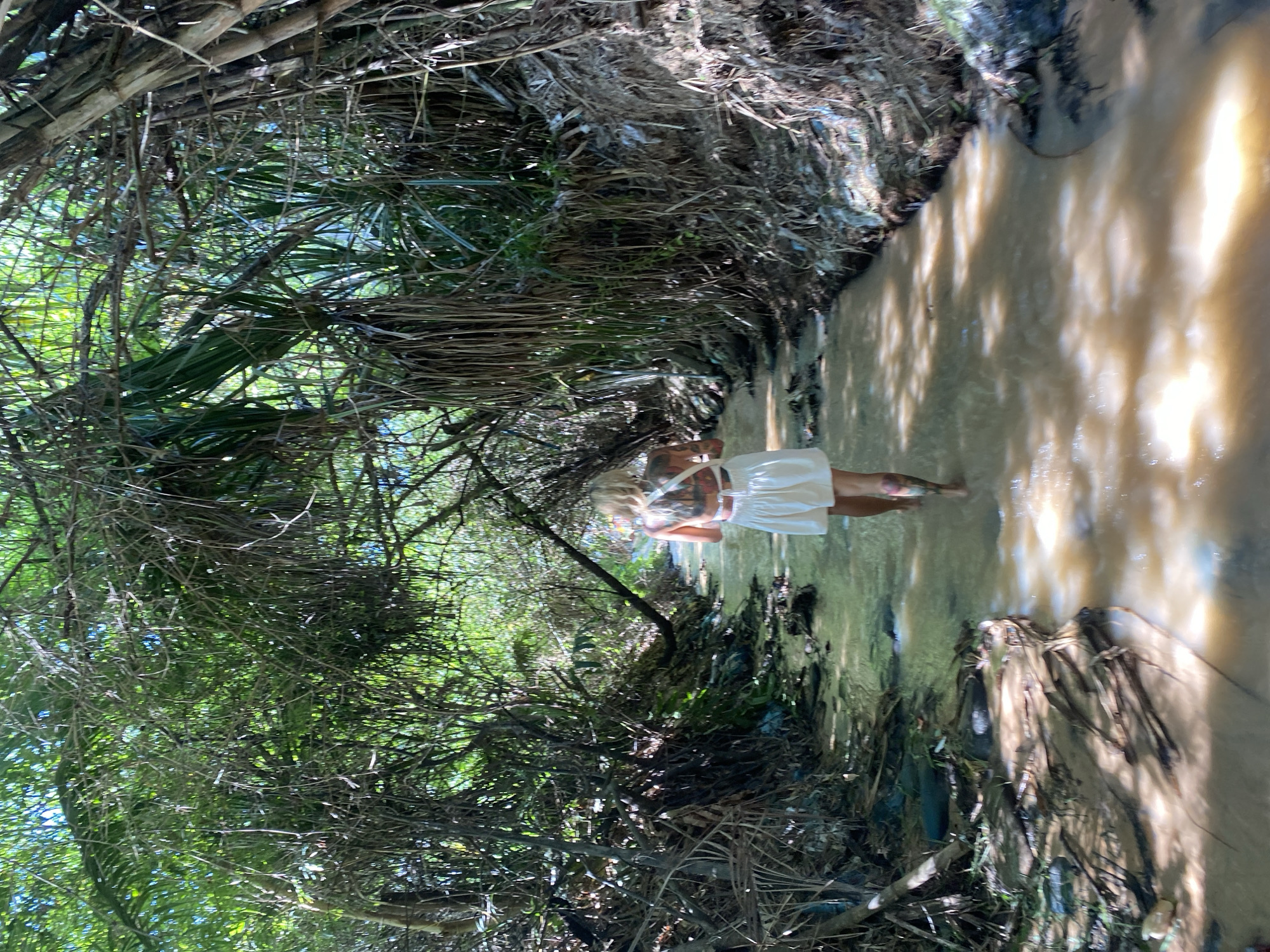 A woman with blond hair and tattoos on her back walks away from the camera through a shallow, muddy stream. She is wearing a white dress. The stream is surrounded by dense vegetation, with trees and branches forming a canopy overhead. The banks of the stream are covered in debris and refuse. The image appears to be taken in a natural, undeveloped area. The specific location of Phu Thuy Ward, Vietnam is not evident from the visual information.