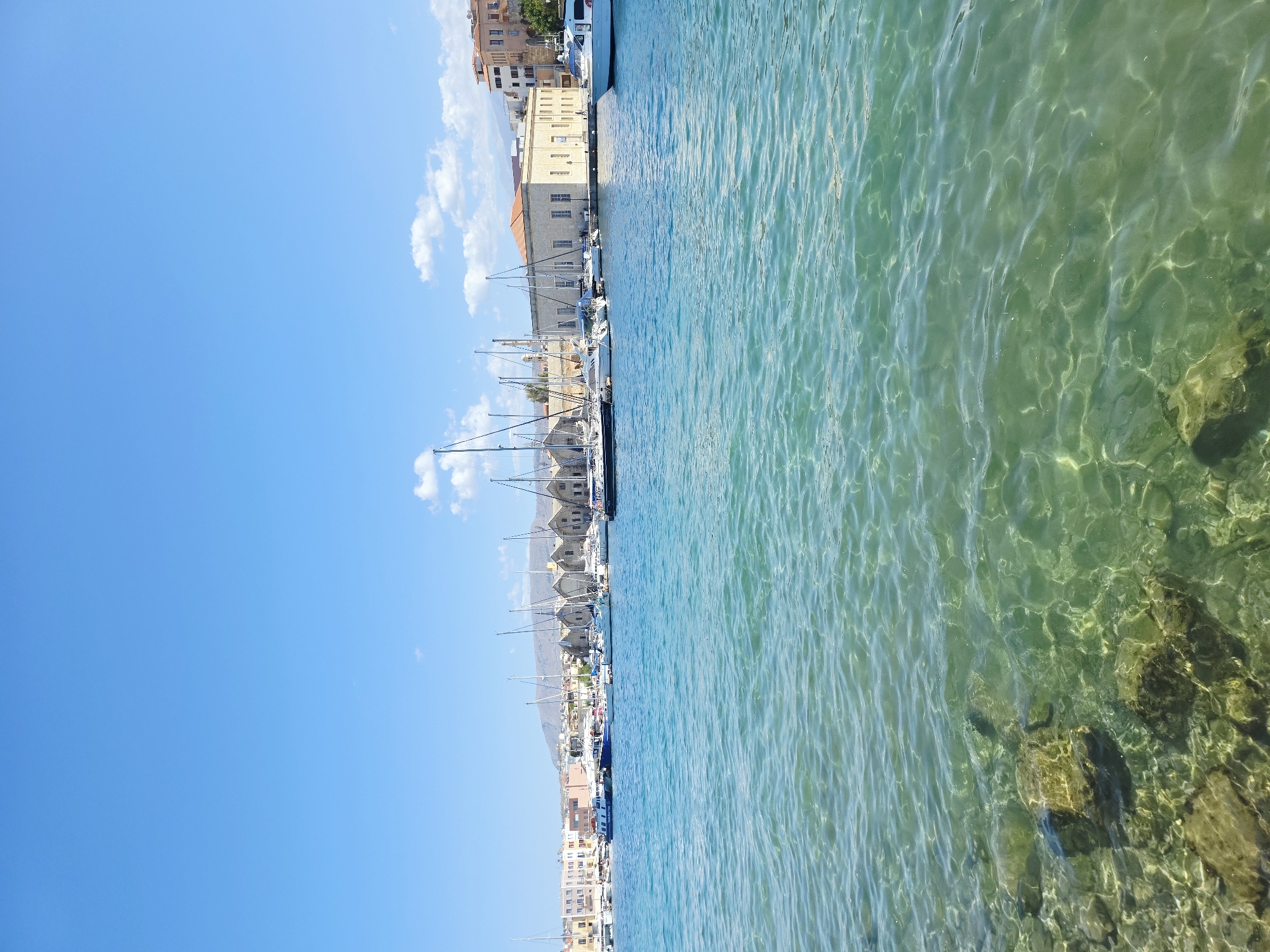 This is a vertical, eye-level shot of the harbor in Chania, Greece. The water in the foreground is a clear, light green with visible rocks and stones on the seabed, and it ripples gently. In the middle ground, a line of sailboats and a few motorboats are docked along the waterfront. The buildings in the background are Mediterranean in style, with terracotta roofs and light-colored walls. A larger, stone building with a light orange roof is prominent, along with other multi-story structures and a distant hillside. The sky is a bright, clear blue with a few wispy white clouds. There are no people visible in the frame, nor any discernable actions or interactions. The scene depicts a peaceful, sunlit day in the harbor.