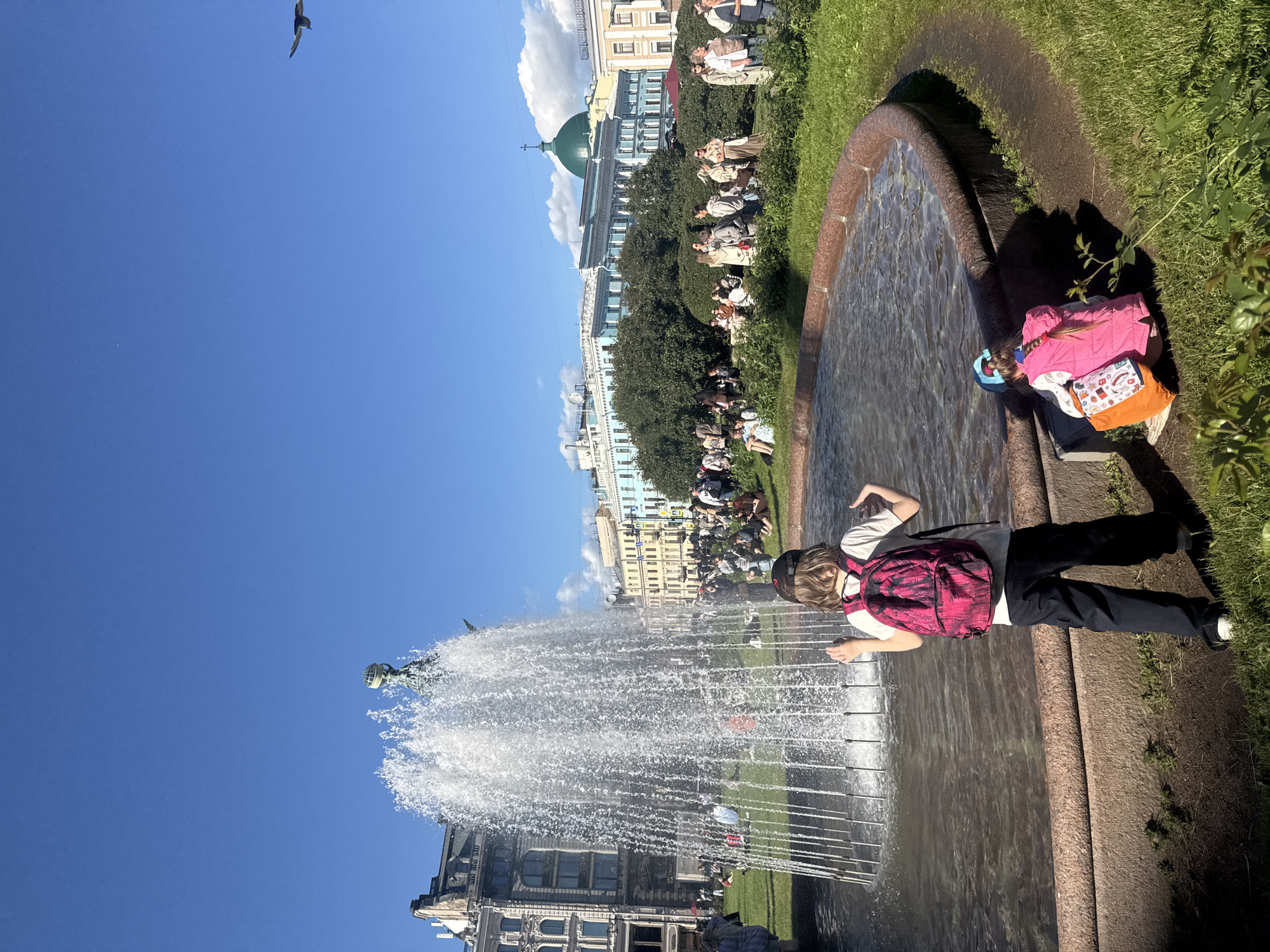 In this sun-drenched scene, people are gathered in a park near a large fountain. A young person with a bright pink backpack stands by the fountain's edge, looking towards the spraying water. Other individuals are scattered across the lush green lawn, some sitting and observing, while others are partially obscured by the dense foliage. In the background, ornate buildings with light-colored facades and green domes line a wide avenue, suggesting an urban setting. The sky is a clear, vibrant blue, with a few wispy clouds contributing to the bright atmosphere. A solitary bird is captured in flight against the expansive sky. The overall impression is a leisurely day in a picturesque city, likely Saint Petersburg, Russia, given the architectural style. The time appears to be daytime, with the sun casting distinct shadows.