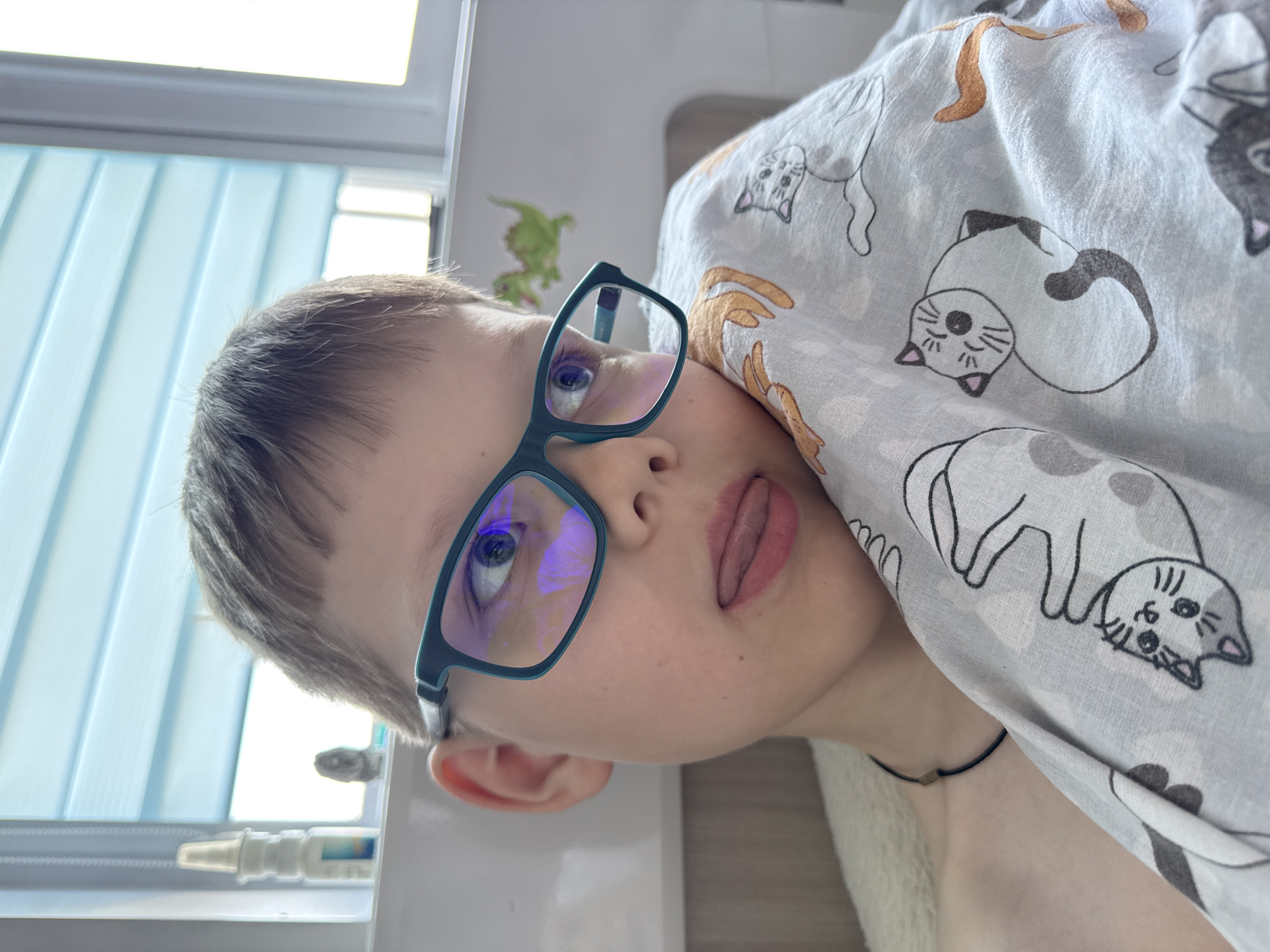A boy with short brown hair and blue-framed glasses is looking at the camera with his tongue sticking out. He is lying down with a light gray blanket patterned with drawings of cats partially covering him. A small black choker is visible around his neck. In the background is a window with light blue blinds. Also visible on the window sill is a bottle of eye drops. A green dinosaur sticker is on the wall. The location may be in or near Gornyi (Dvurechenskoe S/P), Russia, based on the prompt, but this cannot be confirmed from the image alone.