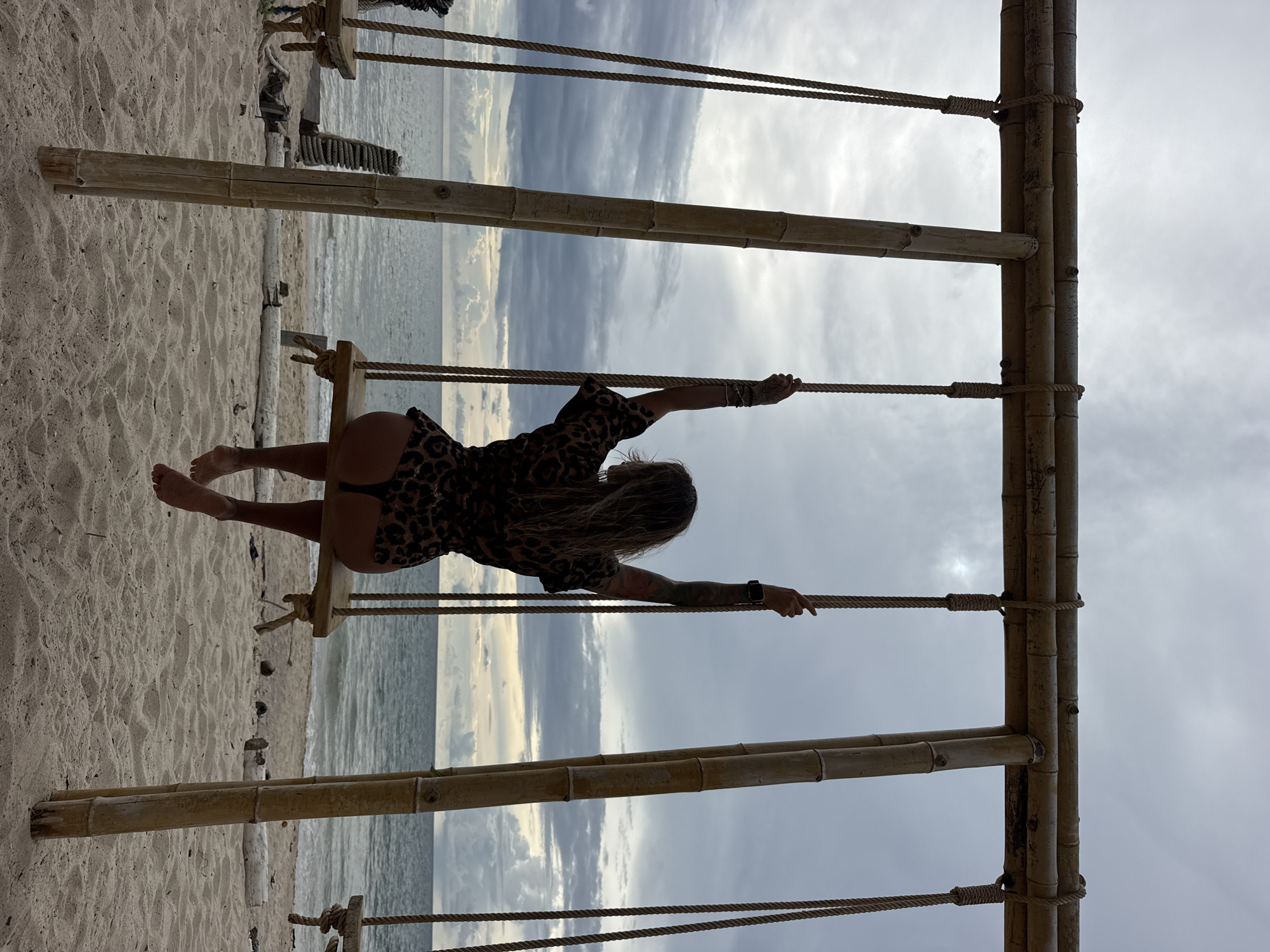 A person sits on a wooden swing, their back to the camera, on a sandy beach. The swing is constructed from bamboo poles and ropes. The person is wearing a leopard-print outfit. The ocean is visible behind them, with a cloudy sky overhead. The location is the city of Choeng Thale, Thailand. The scene appears to be tranquil and serene.