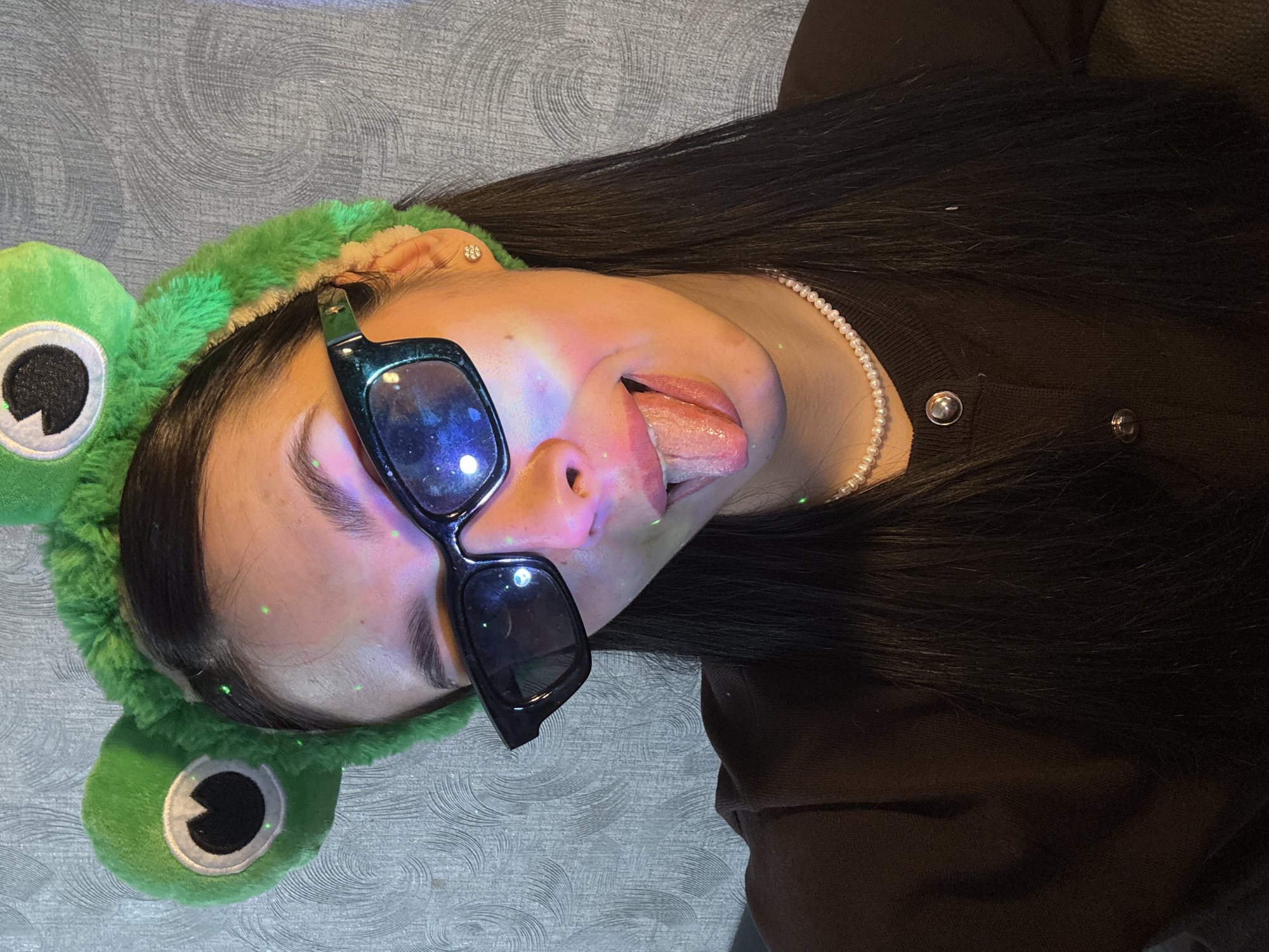 A woman lies on a patterned surface, wearing a green plush frog headband and dark sunglasses. She is wearing a pearl necklace and a dark brown top with silver buttons. She sticks out her tongue. The location is unknown.