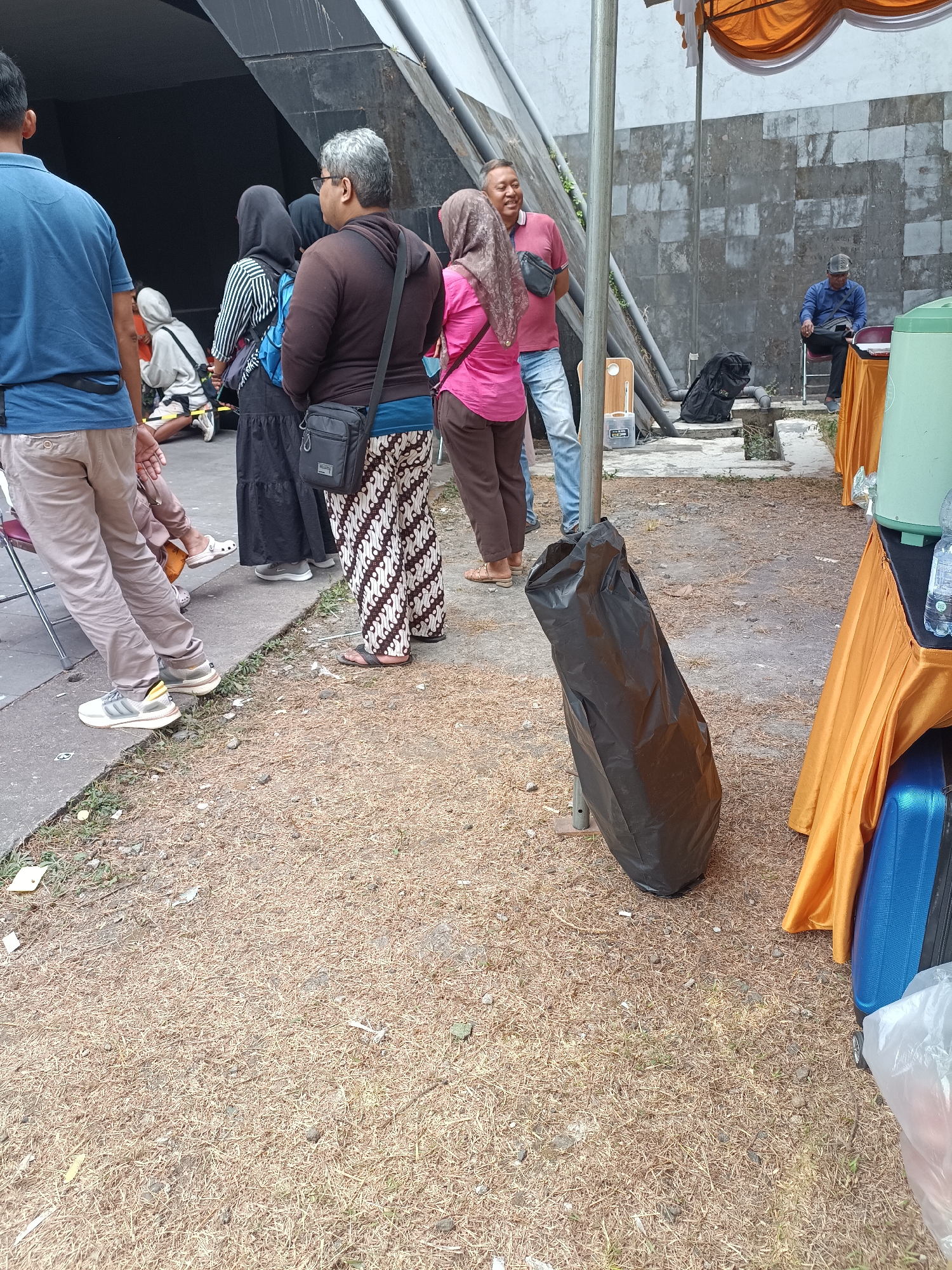 The image depicts a scene in what is described as Hargobinangun, Indonesia. Several individuals are present, primarily in the foreground and midground. A man in a blue polo shirt and tan pants is on the far left, facing away from the camera. Next to him, a person in a grey hooded jacket is kneeling. A group of people are standing together, including a woman in a black hijab and striped shirt, a person in a brown hoodie with a black crossbody bag, and a woman in a pink shirt and headscarf. To the right, a man in a red polo shirt and blue jeans is smiling. Further to the right, a man in a blue shirt and cap is seated on a chair at a table covered with an orange cloth.
The environment appears to be outdoors, with a paved area on the left transitioning to a ground covered in dried pine needles and some scattered debris. A large black trash bag is leaning against a metal pole. Behind the people, a dark opening suggests an underpass or entrance. A light-colored wall with a textured surface forms the background on the right. Orange fabric drapes are visible overhead and to the right, covering a large green water dispenser. A blue suitcase and a white plastic bag are also visible on the ground to the right. The overall impression is of a public gathering or waiting area.