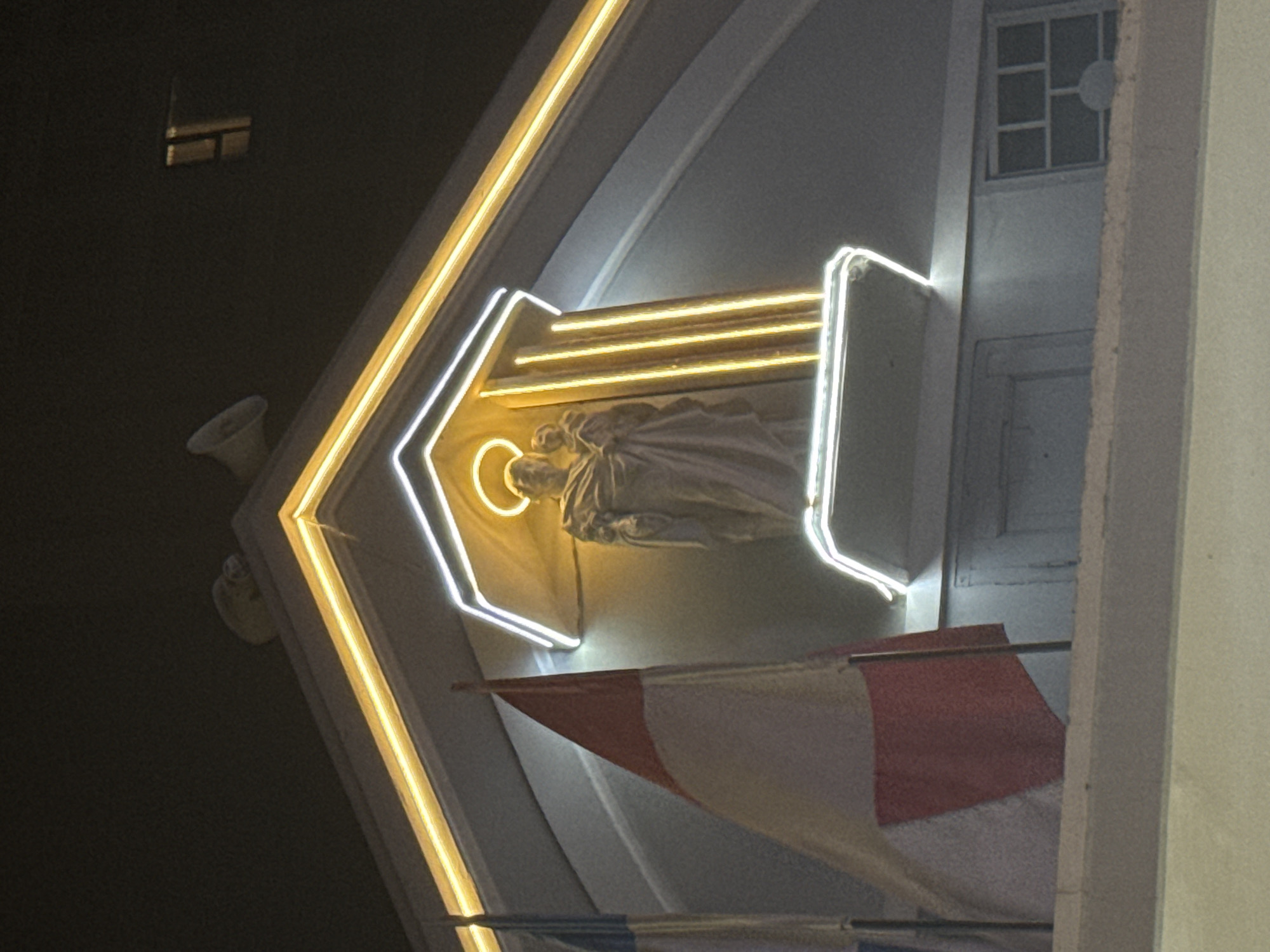 A nighttime, low-angle shot shows a building facade in Nha Trang Ward, Vietnam. A statue is lit by white and yellow neon lights inside an alcove. A red and white flag hangs below the statue. A speaker is visible to the left. A white grid window and part of an adjacent building are on the right.