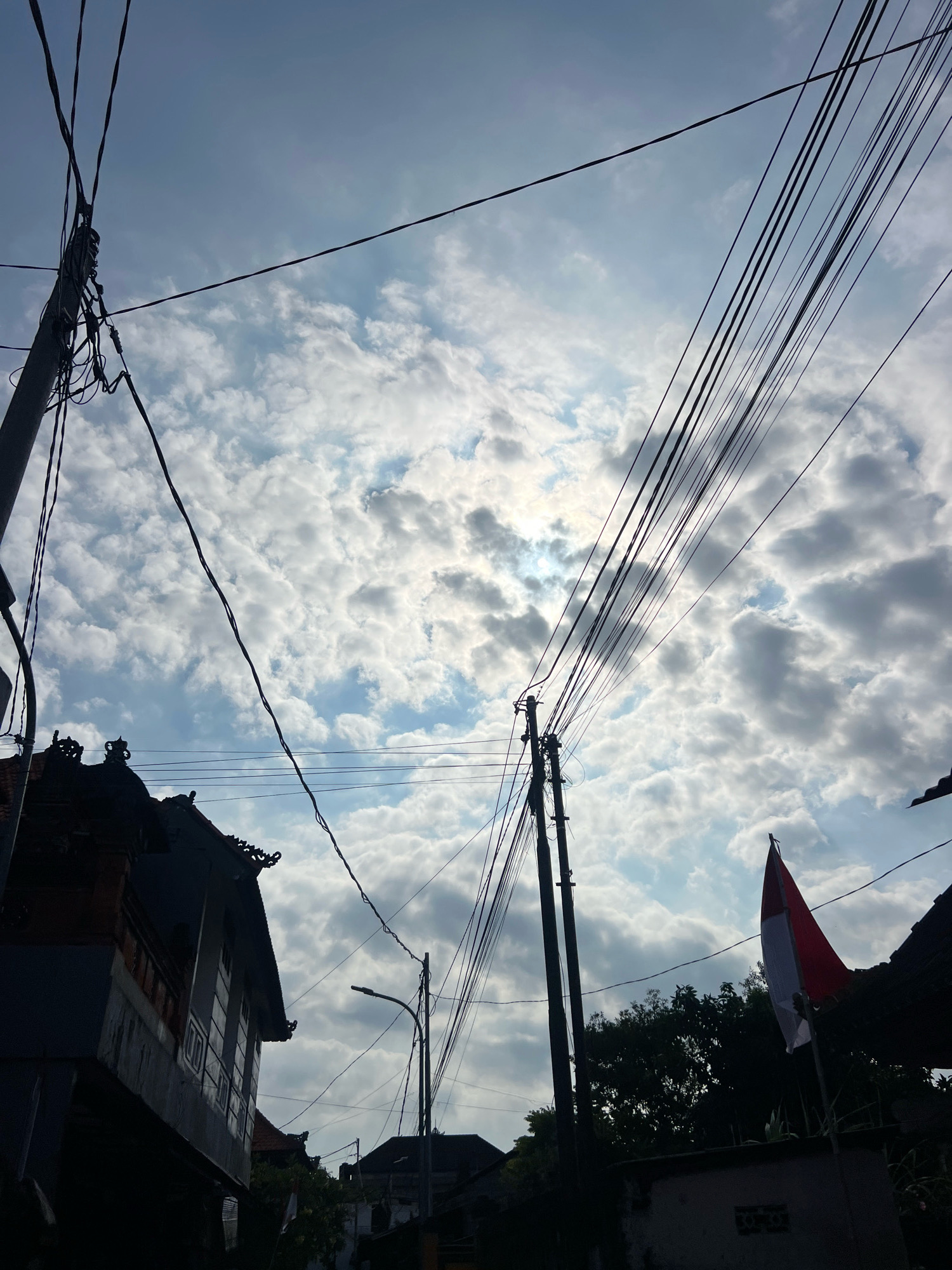 This is a medium-shot, low-angle photograph taken outdoors in Kuta, Indonesia. The scene captures a cloudy sky with the sun partially obscured by clouds. In the foreground, there is a silhouette of a building with traditional Balinese architectural elements, including intricate roof decorations. Several utility poles with numerous overhead wires stretch across the frame, creating a dense network against the sky. To the right, a red and white flag, likely the Indonesian flag, is mounted on a pole. Silhouetted trees and other buildings are visible in the background, suggesting a residential or urban area. The lighting indicates it is either early morning or late afternoon. There are no visible people or animals in the frame.
