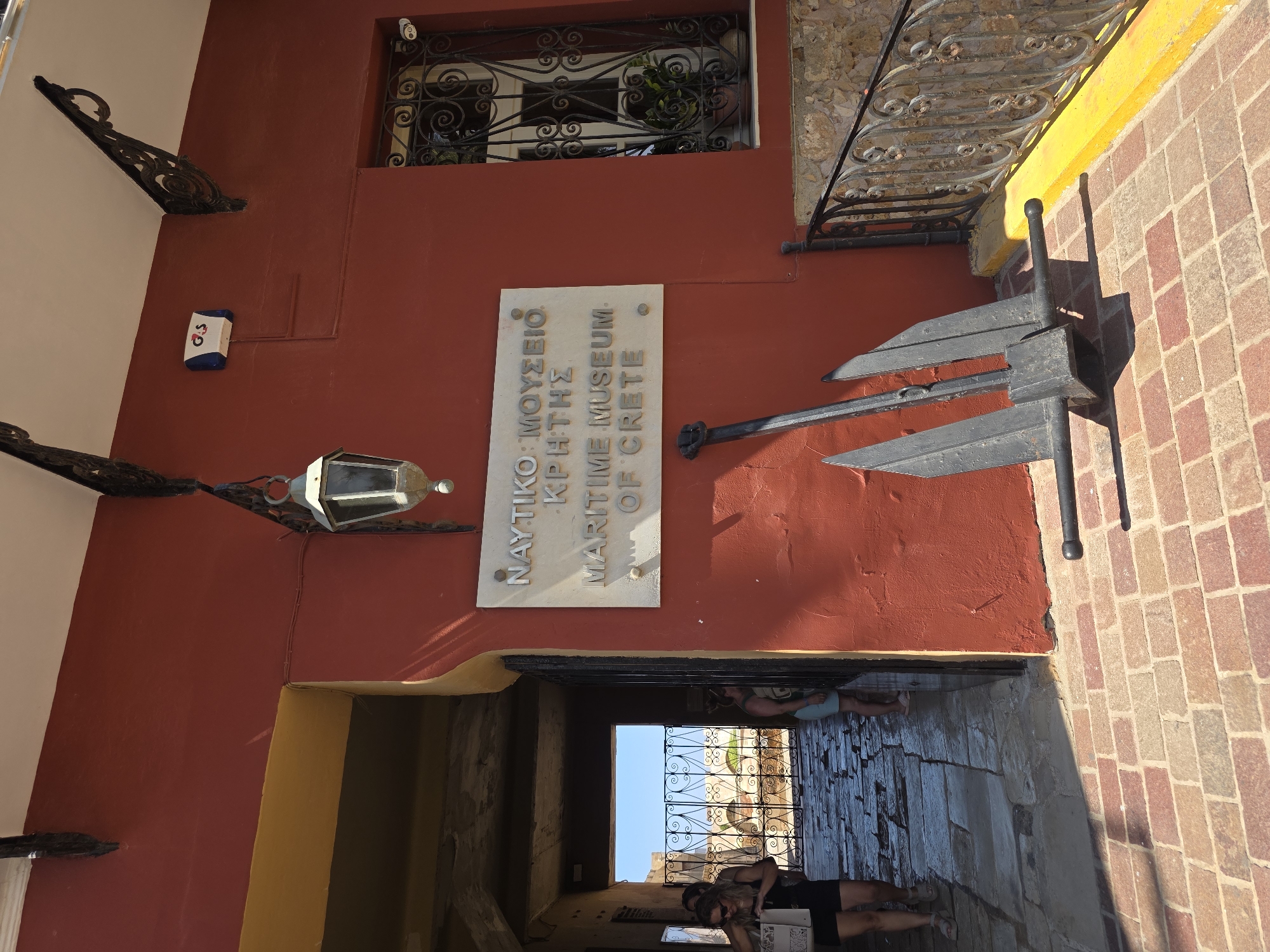 The image shows the entrance to the Maritime Museum of Crete in Chania, Greece. The museum entrance is a red building with a yellow archway. A sign above the entrance reads "NAUTIKO MOYΣEIO KPHTHΣ MARITIME MUSEUM OF CRETE". A large black anchor is placed in front of the museum. In the background, through the archway, there are glimpses of stone structures and decorative iron gates. Two women are seen walking through the archway. The ground is paved with stone tiles and bricks, and there is a black wrought-iron railing on the right side of the image. The sky is clear and blue, indicating a sunny day.