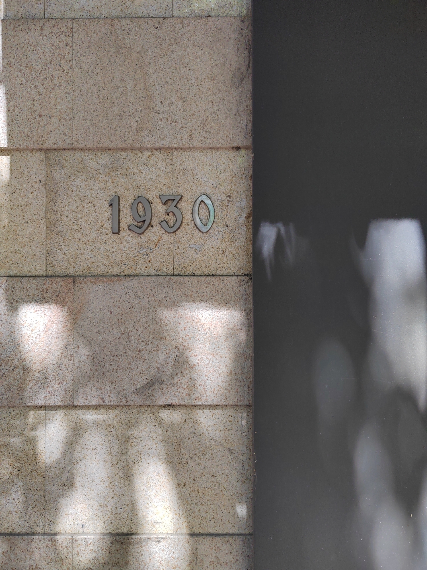 The image shows a section of a light brown, speckled stone wall with the number "1930" affixed to it in metal numerals. A dark, solid, non-reflective surface adjoins the stone wall on the right. There are shadows cast on the stone wall, suggesting the presence of foliage or other structures blocking direct sunlight. The location is likely a building exterior in Podgorica, Montenegro, due to the presence of a building with the number 1930.